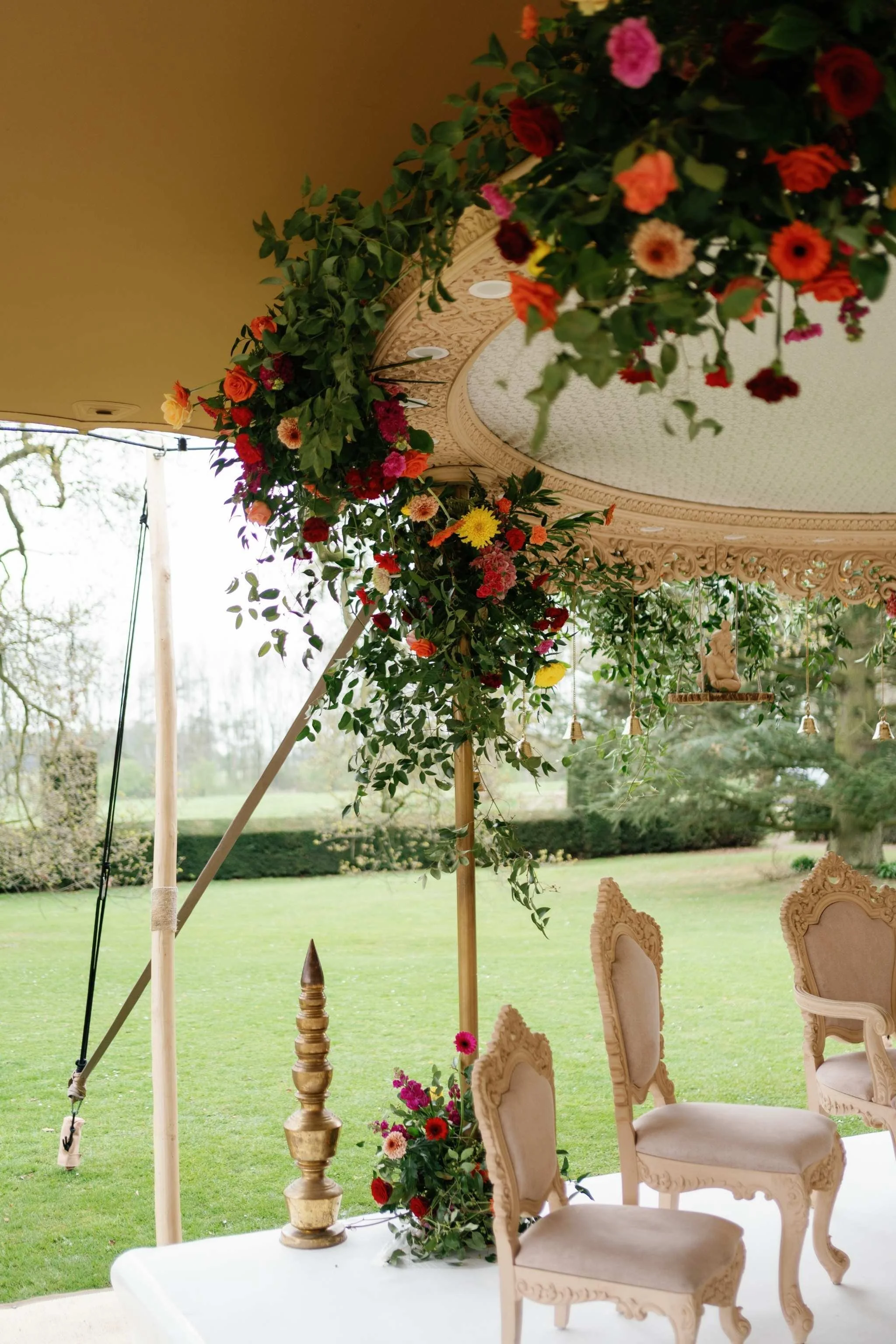 Elegant white chairs with ornate carvings arranged in a row outdoors under a decorated canopy with floral garlands and a table with a floral centerpiece. Green grass and trees are visible in the background.