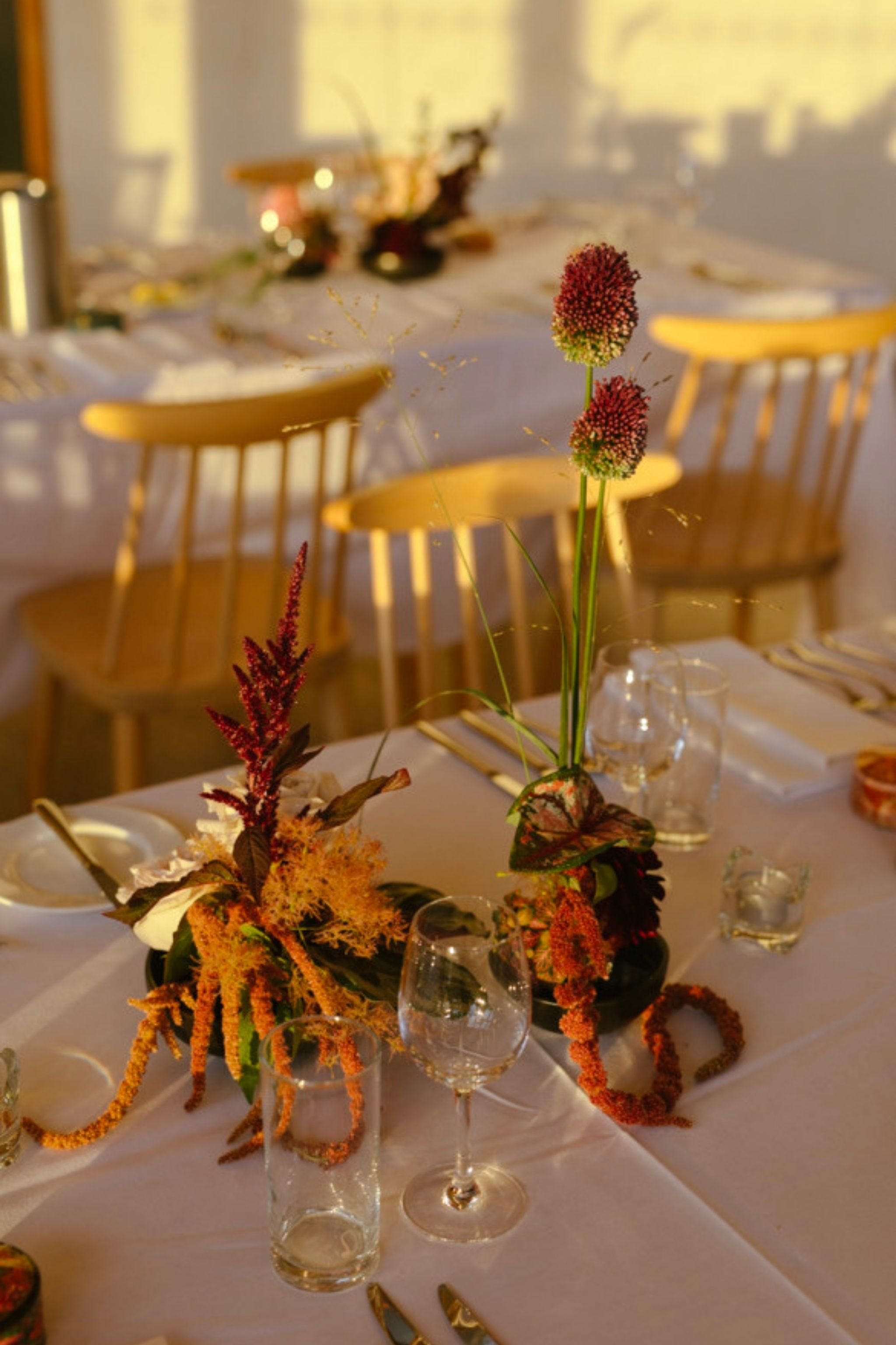A table setting with floral centerpieces, glasses, and cutlery, illuminated by warm sunlight, with wooden chairs in the background.