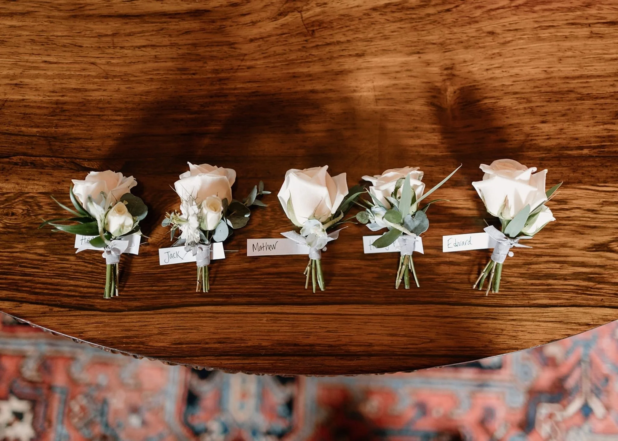 Row of small white floral boutonnieres with green leaves, each labeled with a name, placed on a wooden surface.