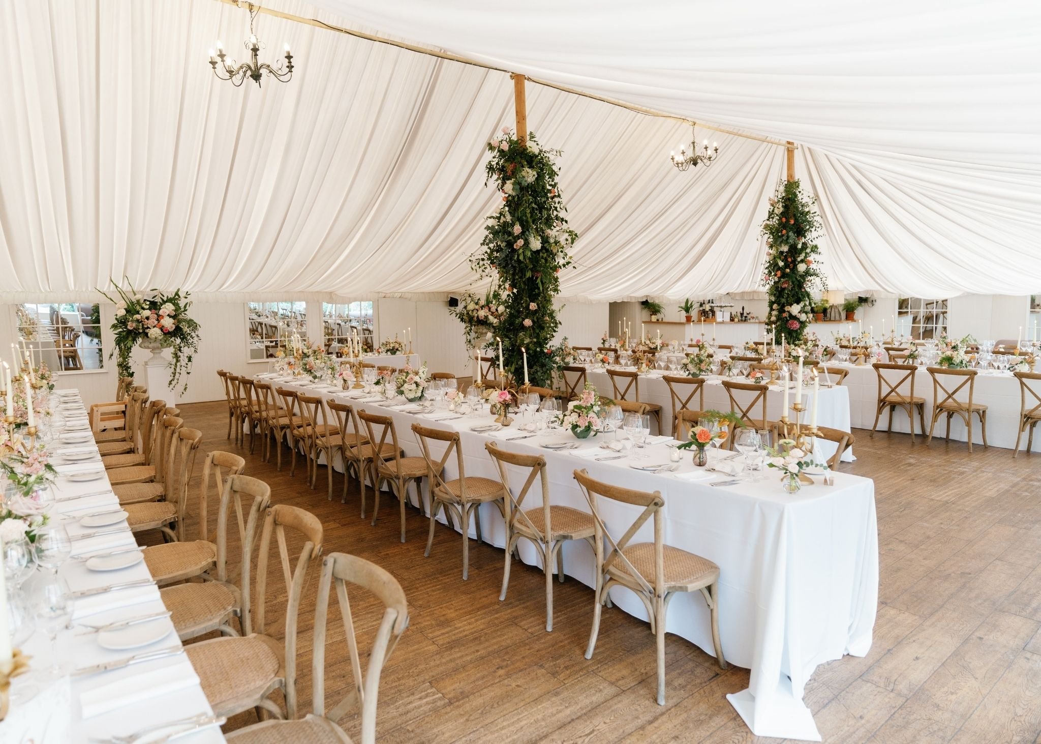 A decorated event tent with long tables set for a celebration, featuring floral centerpieces, white tablecloths, and candle holders under a white draped ceiling with chandeliers and hanging floral arrangements.