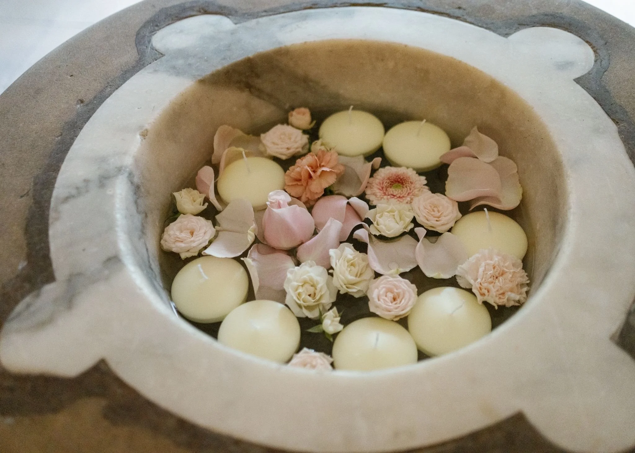 A stone candle holder filled with white candles, pink and white flower petals, and small decorative flowers.