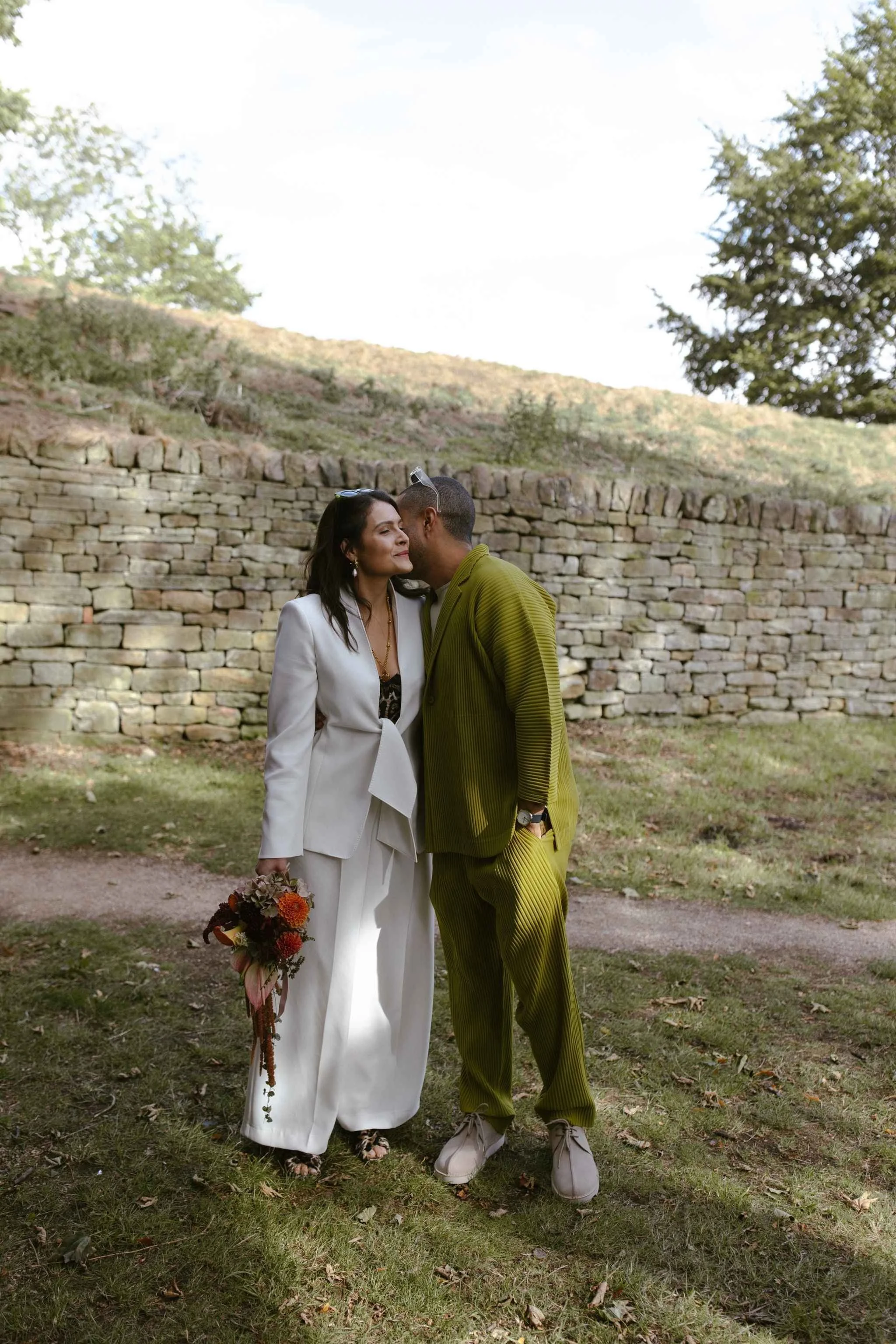 A couple dressed in wedding attire sharing an intimate moment outdoors in front of a stone wall and green hillside, with trees and a clear sky in the background.