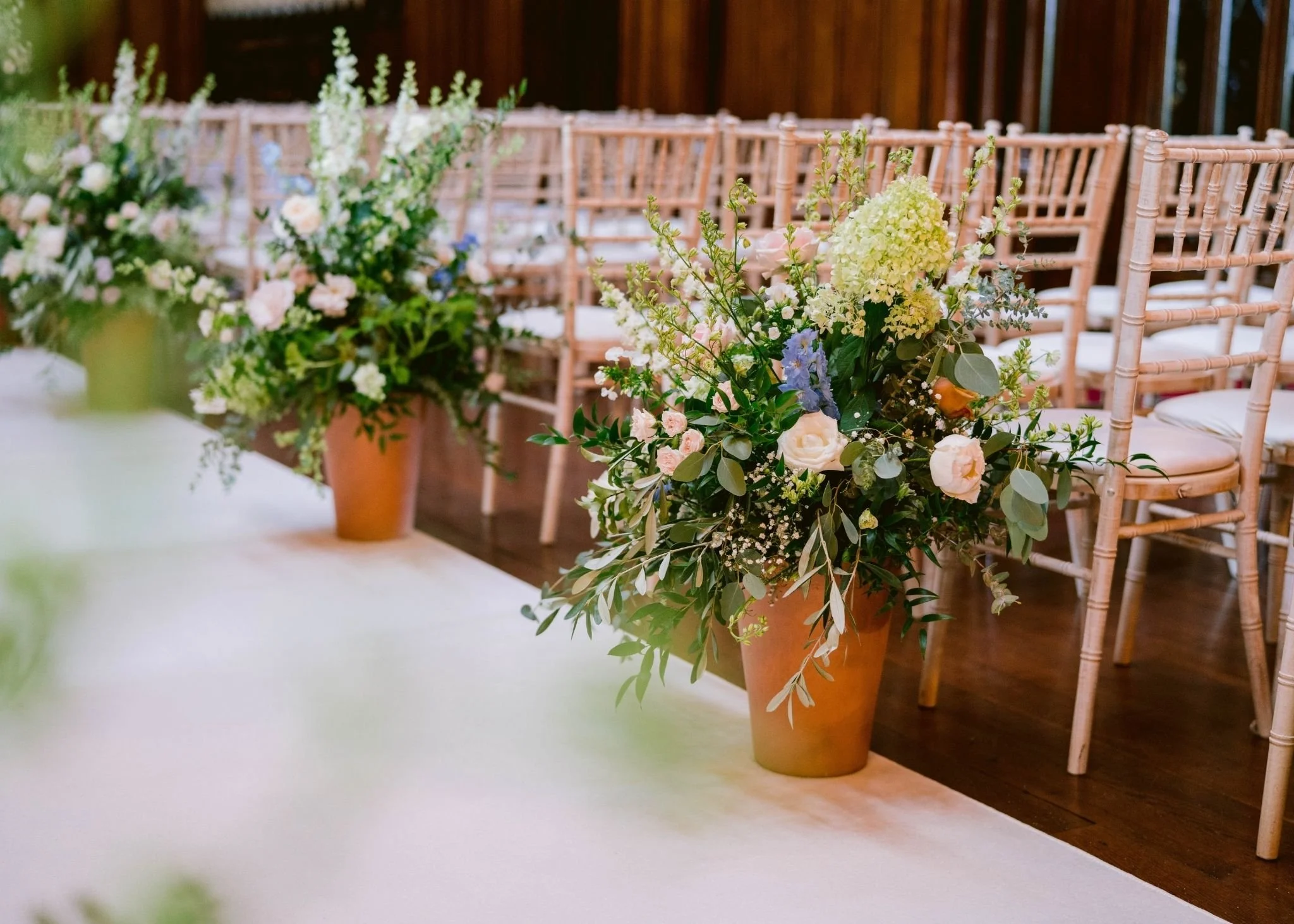 Flower arrangements in terracotta pots with white, pink, and light green blossoms placed along a tableside at a wedding or event with rows of wooden chairs in the background.