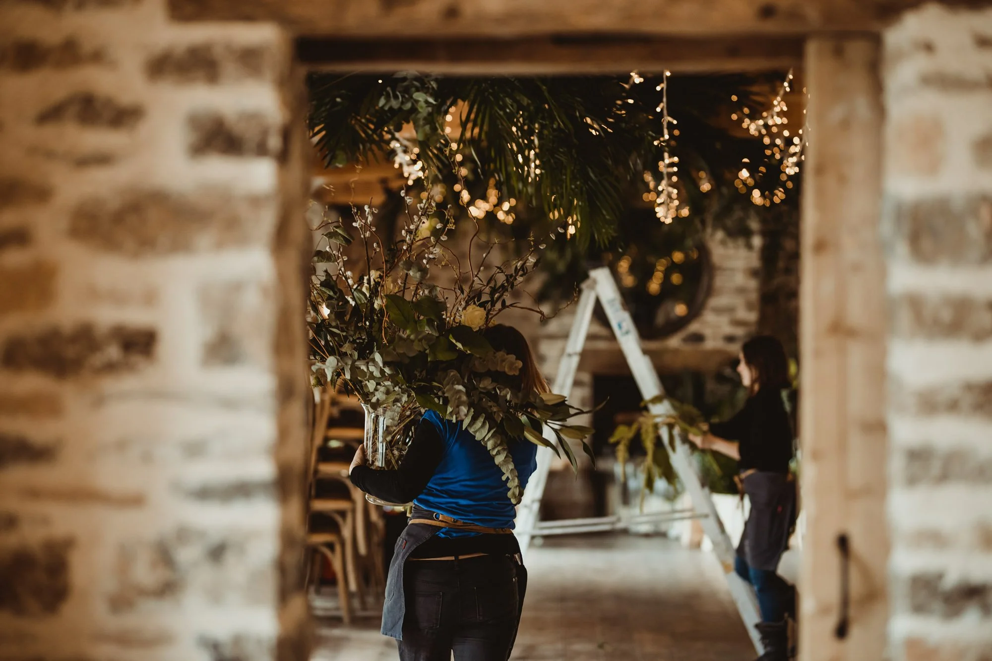 Two people decorating with plants and fairy lights inside a rustic brick building.