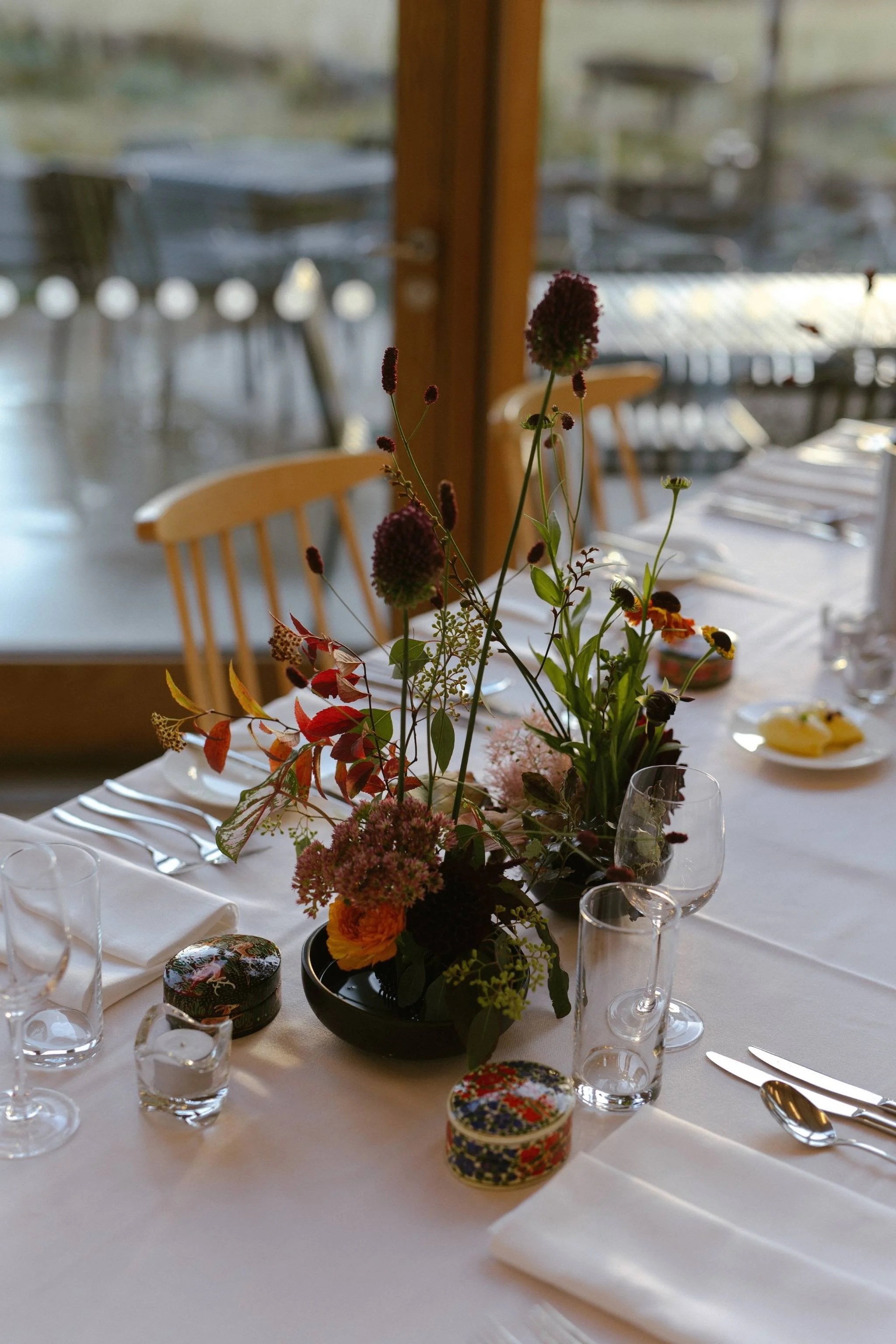 A floral centerpiece with various flowers in a black vase on a white tablecloth, surrounded by clear glassware, plates, and silverware at a dining table set for a special occasion.