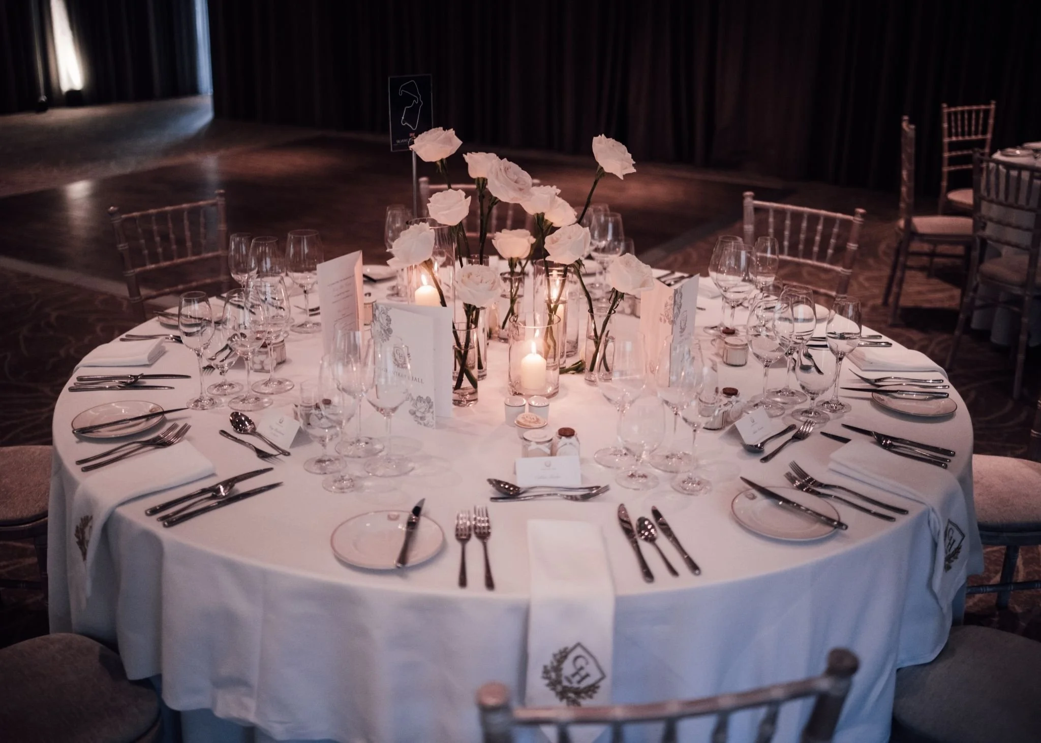 Elegant round banquet table decorated with white flowers in vases, candles, and table settings including glasses, plates, and silverware, set for an event in a dimly lit room.