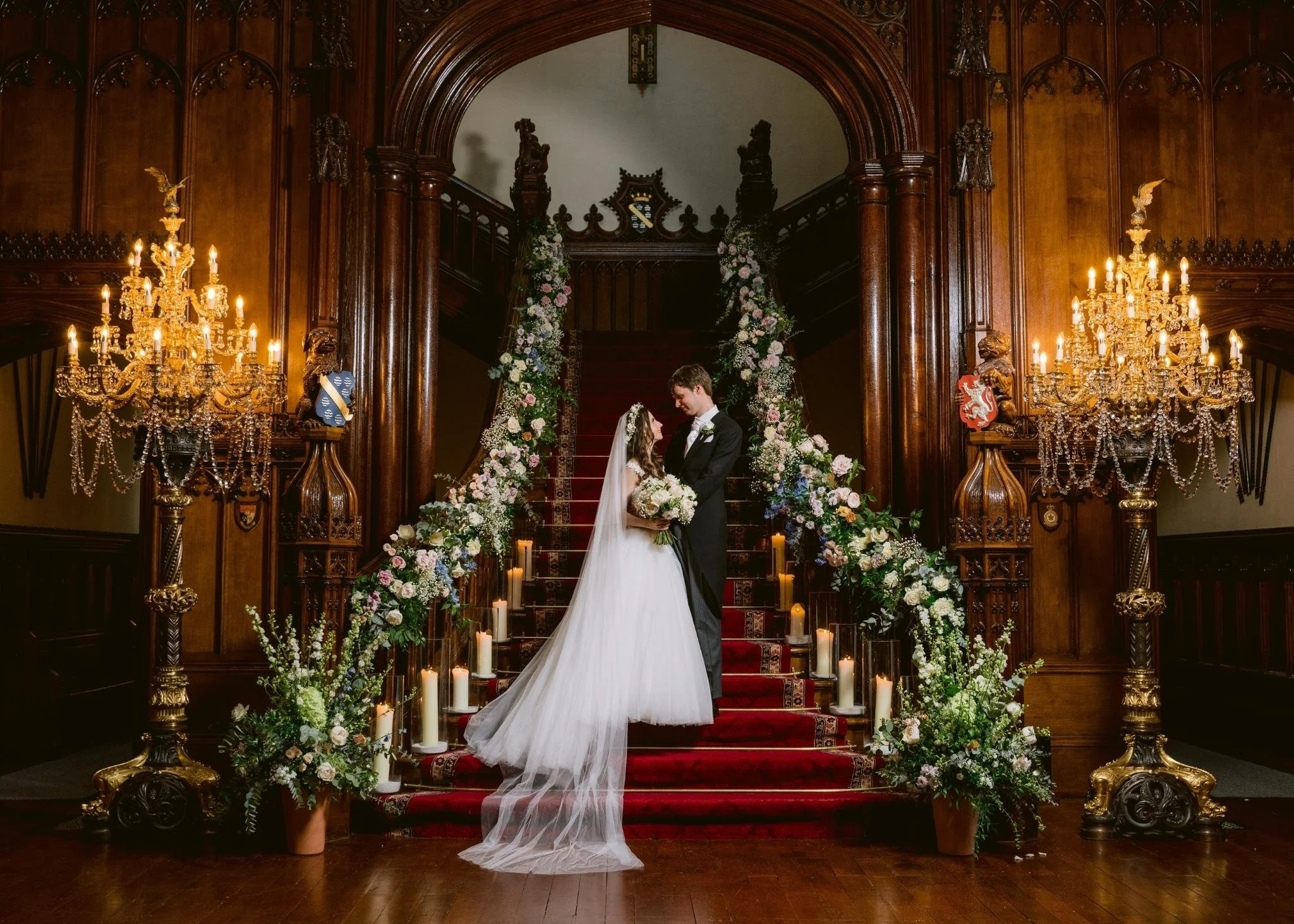 A bride and groom standing on a grand staircase decorated with flowers and candles, inside a richly wood-paneled room with elaborate chandeliers and antique carvings.