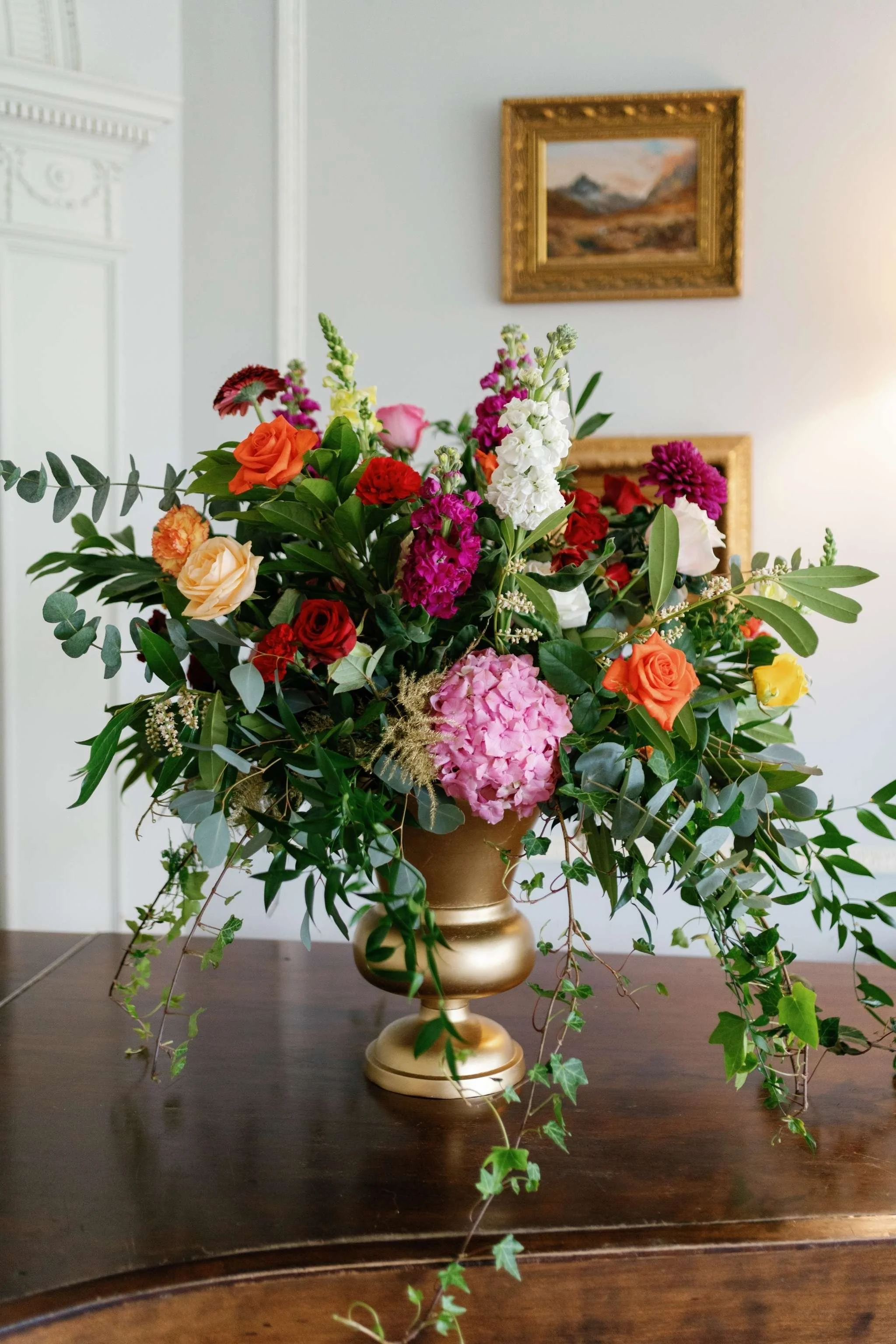 A tall beige vase with a bouquet of multicolored flowers and greenery on a dark wooden table in a room with white walls, framed landscape paintings, and a light source to the right.