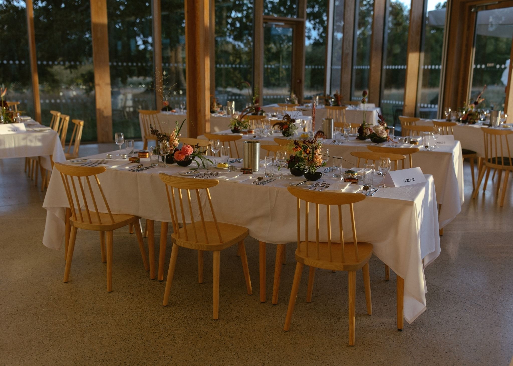 Interior of a restaurant or banquet hall with round tables covered in white tablecloths, decorated with flower arrangements, surrounded by wooden chairs, and set with glassware and cutlery. Large windows in the background reveal trees and outdoor sce