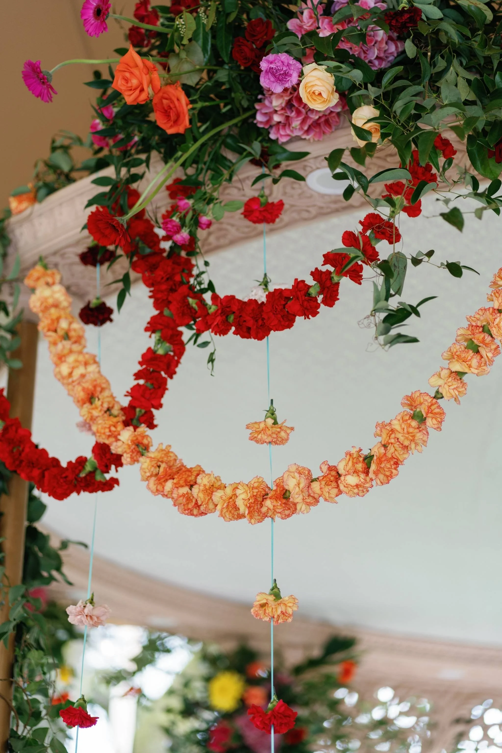 Colorful flower garlands hanging in a floral display. The garlands are made of various red, orange, pink, and peach flowers, suspended on thin strings in a curved arrangement. Green leaves are also visible among the flowers.