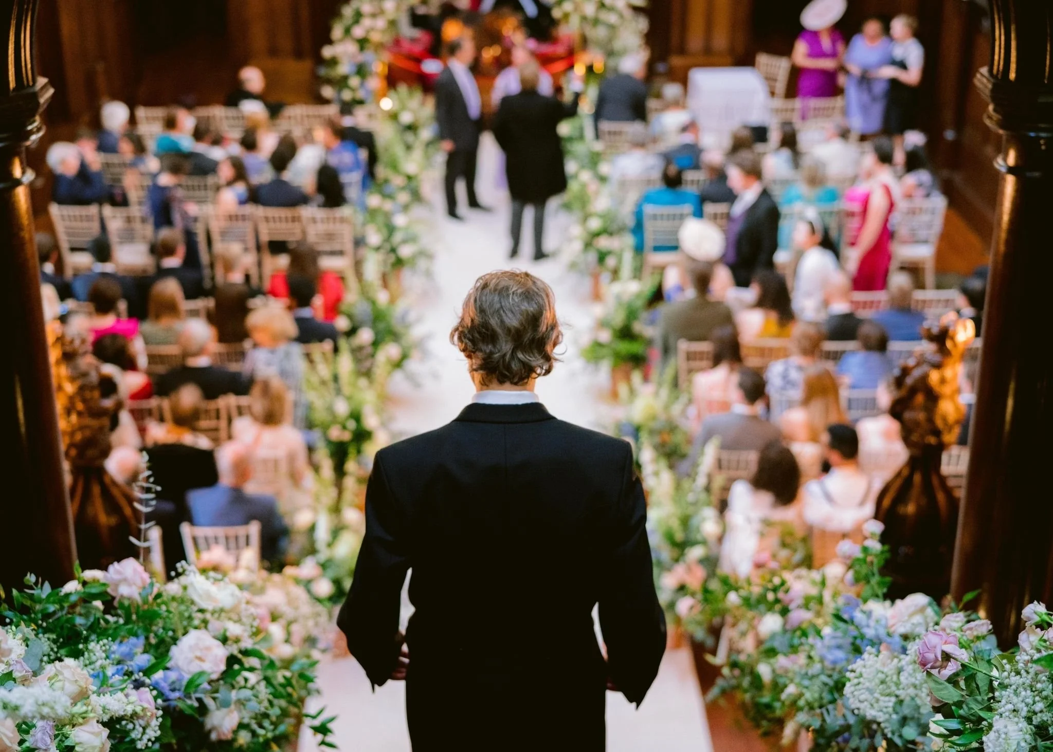 A man in a black suit, seen from behind, walking down an aisle decorated with flowers at a wedding ceremony with numerous seated guests and a floral backdrop at the front.