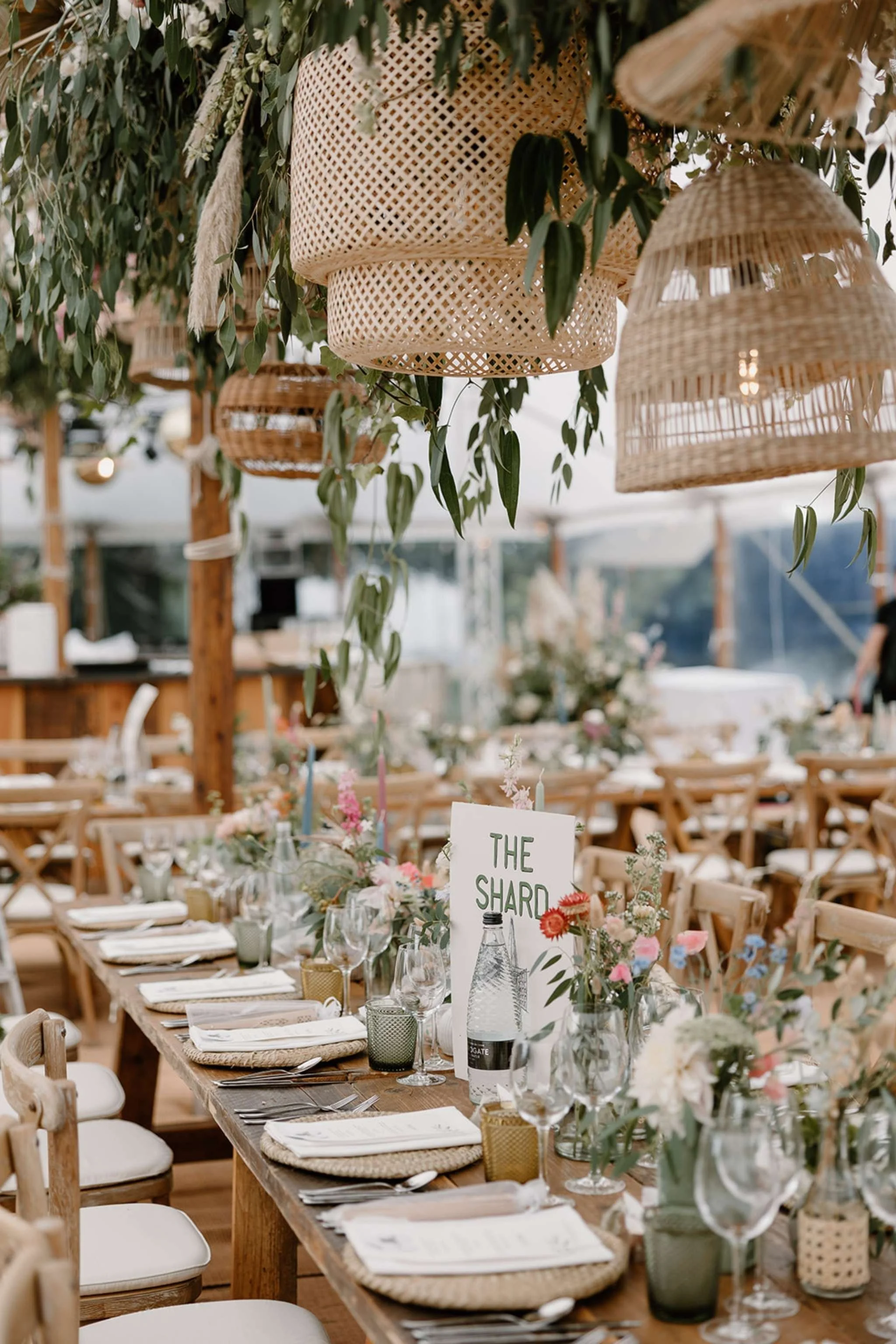 A long wooden table set for a wedding reception with white chairs, floral centerpieces, and hanging woven lanterns, decorated with greenery and soft lighting.