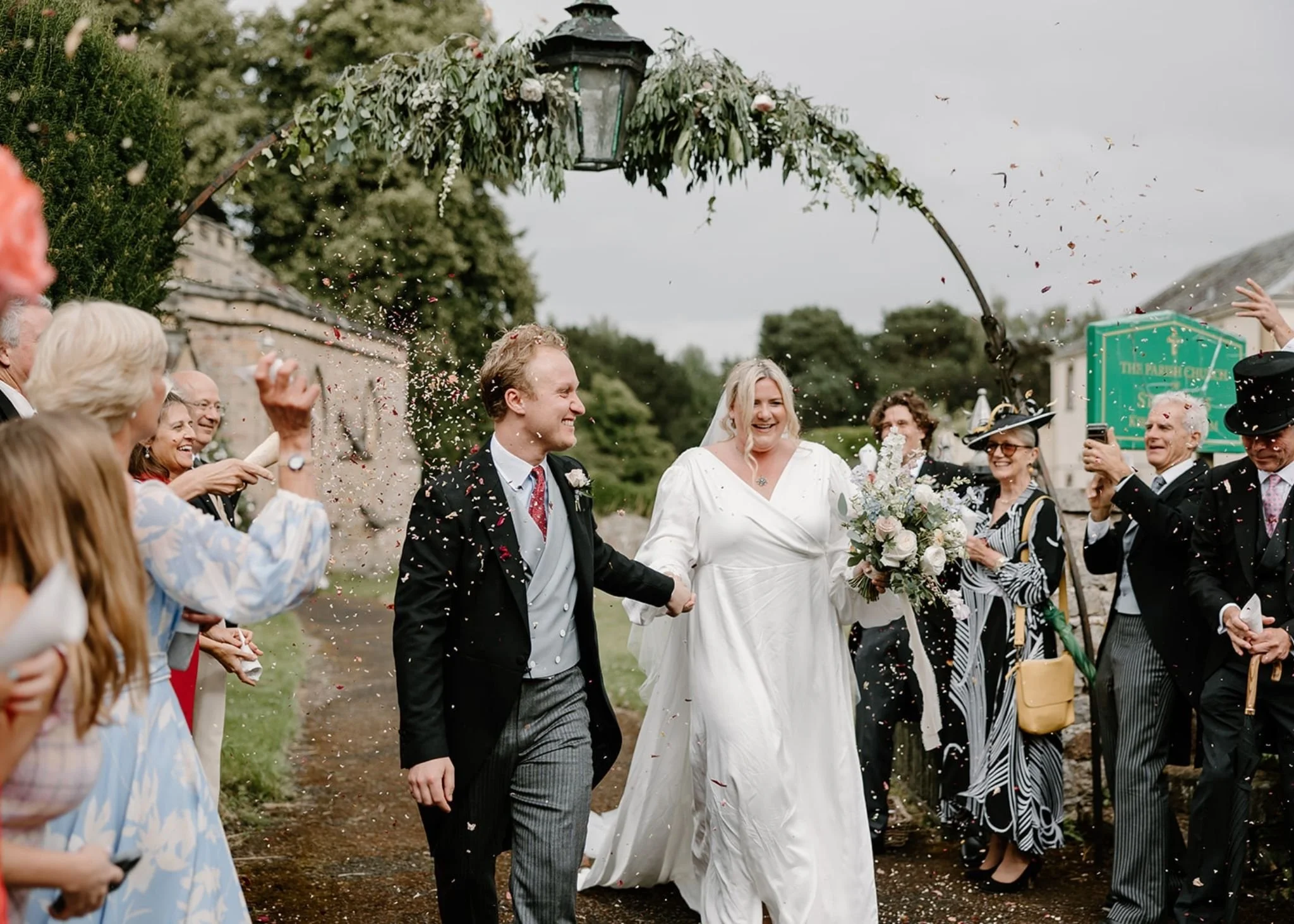 A bride and groom walking outdoors surrounded by family and friends throwing flower petals, celebrating a wedding.