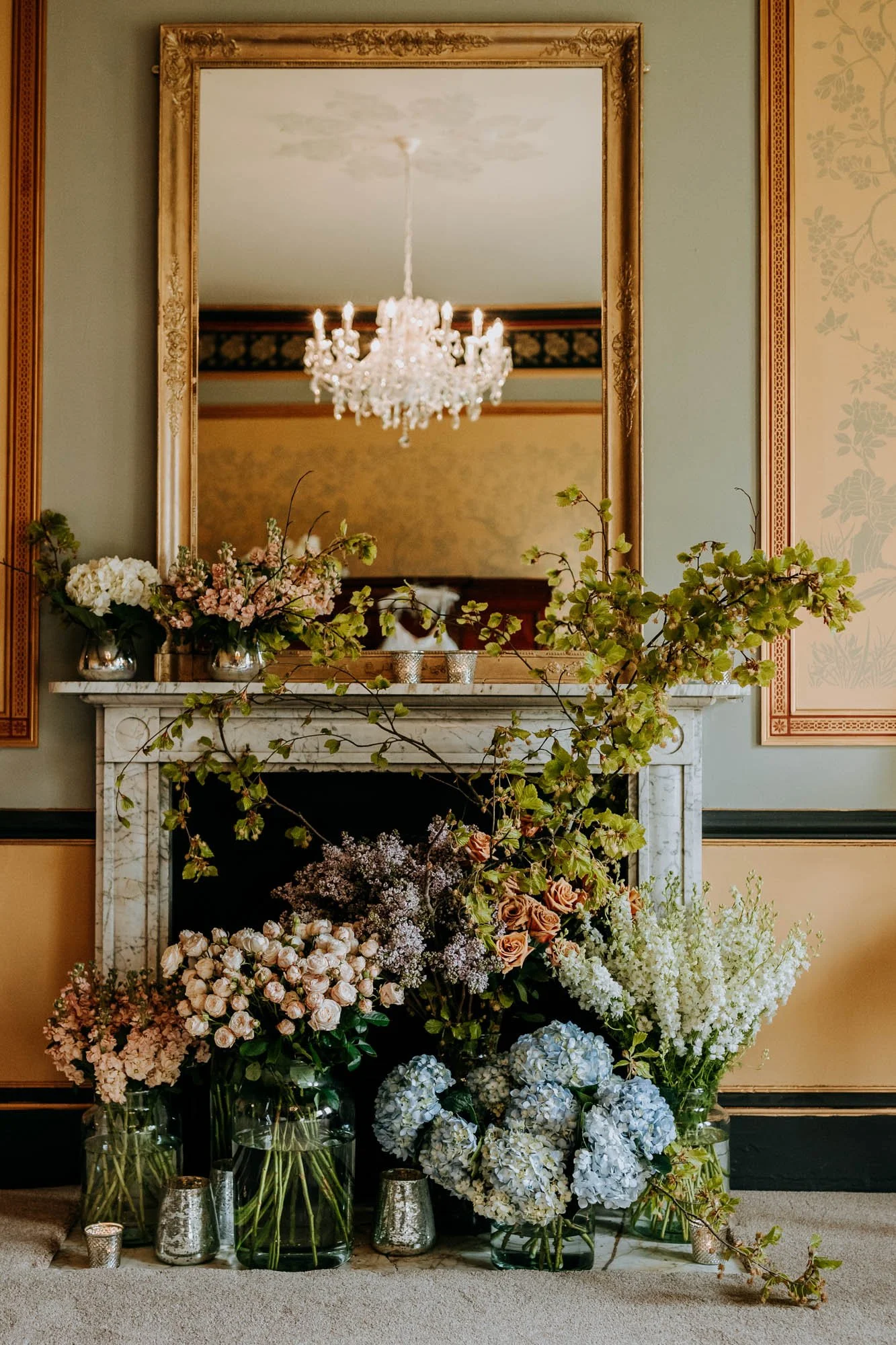 Elegant room with a marble fireplace decorated with various vases of flowers, a large mirror above the mantel reflecting a chandelier, and wall panels with decorative trim.