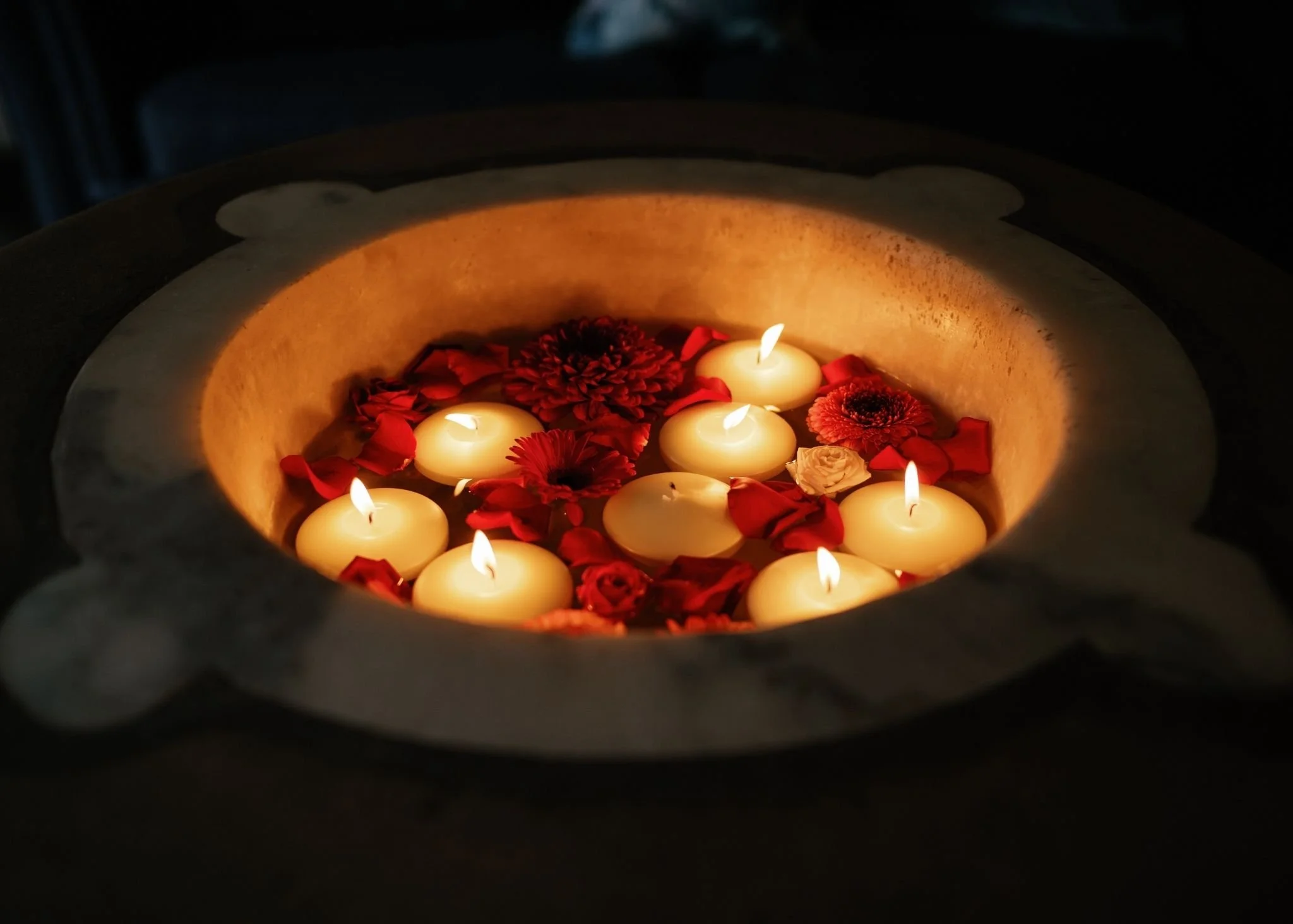 Several lit candles floating in a bowl filled with rose petals and flowers, creating a warm, soft glow.