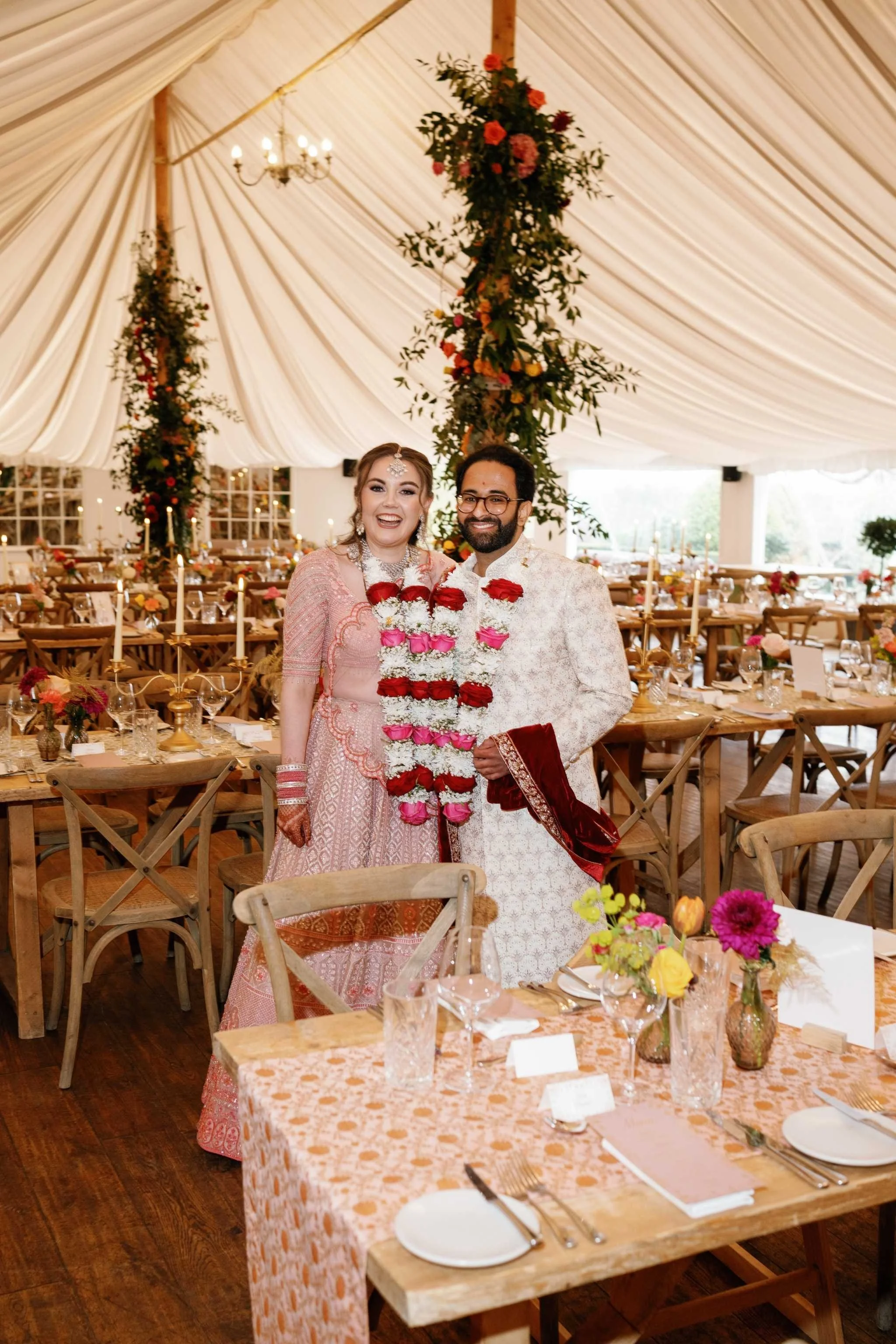 A bride and groom at a wedding reception, standing together smiling, with decorated tables and floral arrangements in a tented venue.