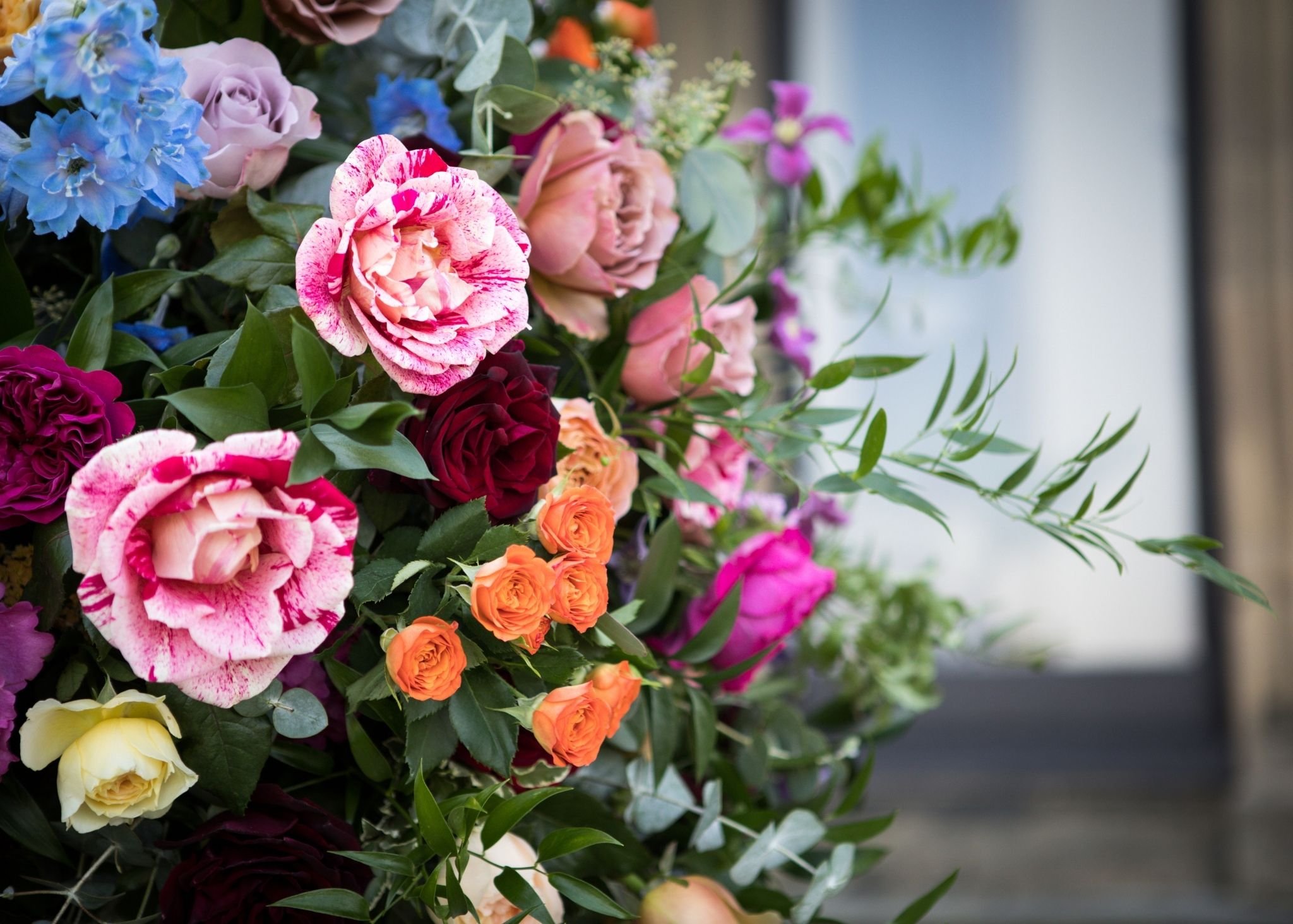 A large bouquet of multicolored flowers, including pink and white speckled roses, orange roses, purple and blue blossoms, and greenery.