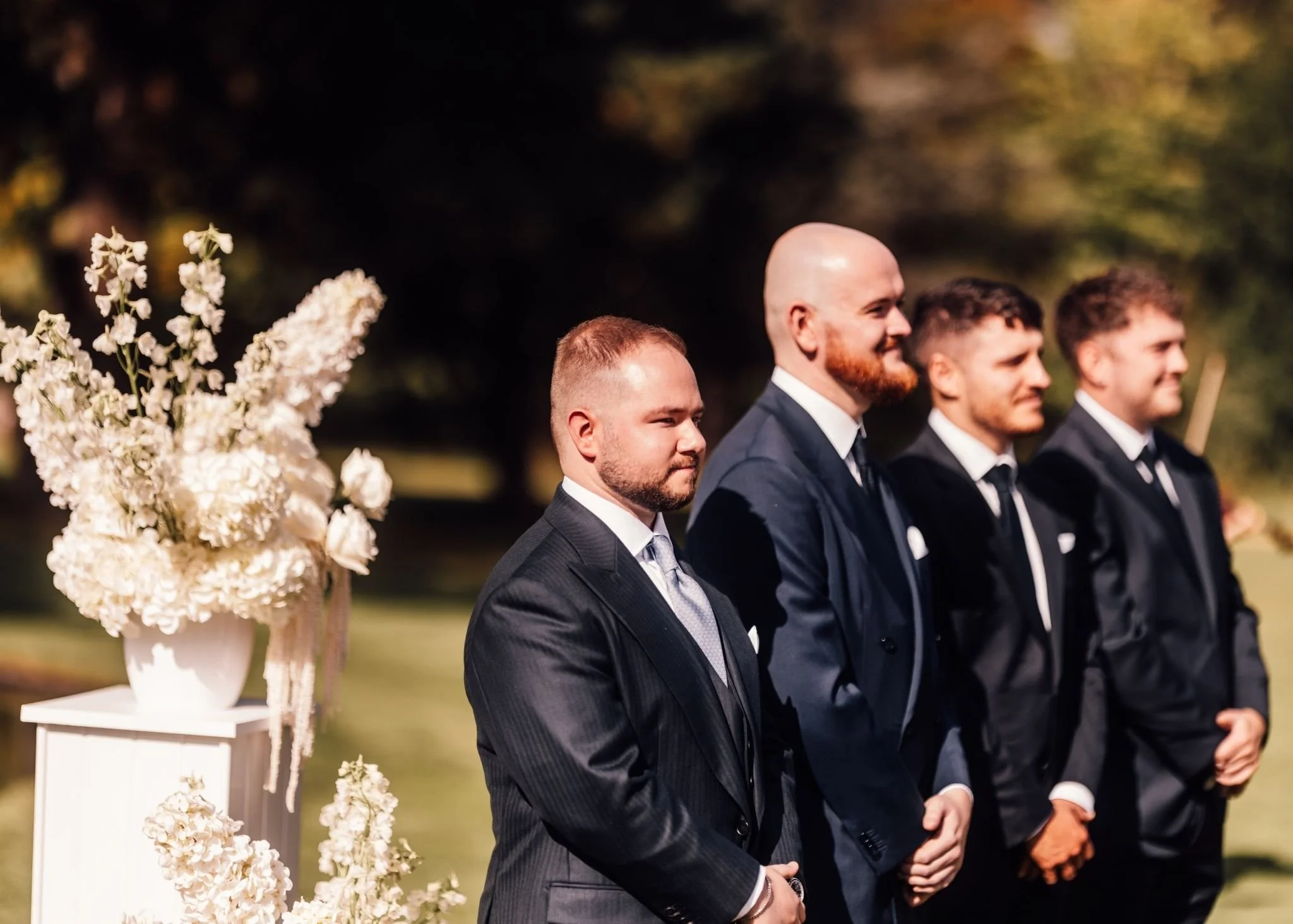 A wedding ceremony scene with four men dressed in dark suits, standing side by side outdoors with trees in the background. A white floral arrangement in a vase is visible on a table to the left.