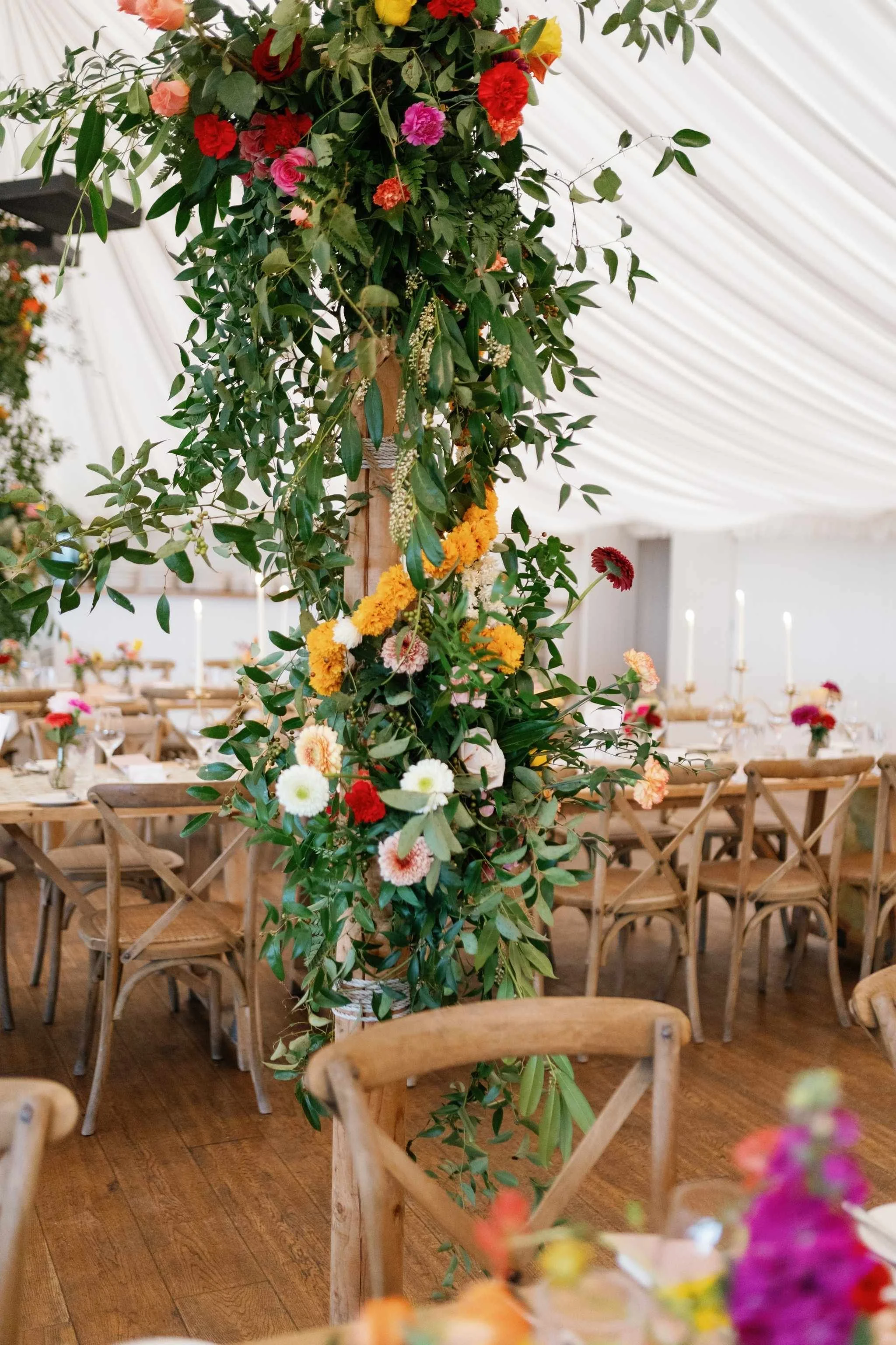A tall floral arrangement with various colorful flowers and green leaves on a wooden column, in a decorated event space with wooden chairs and tables set for a gathering.