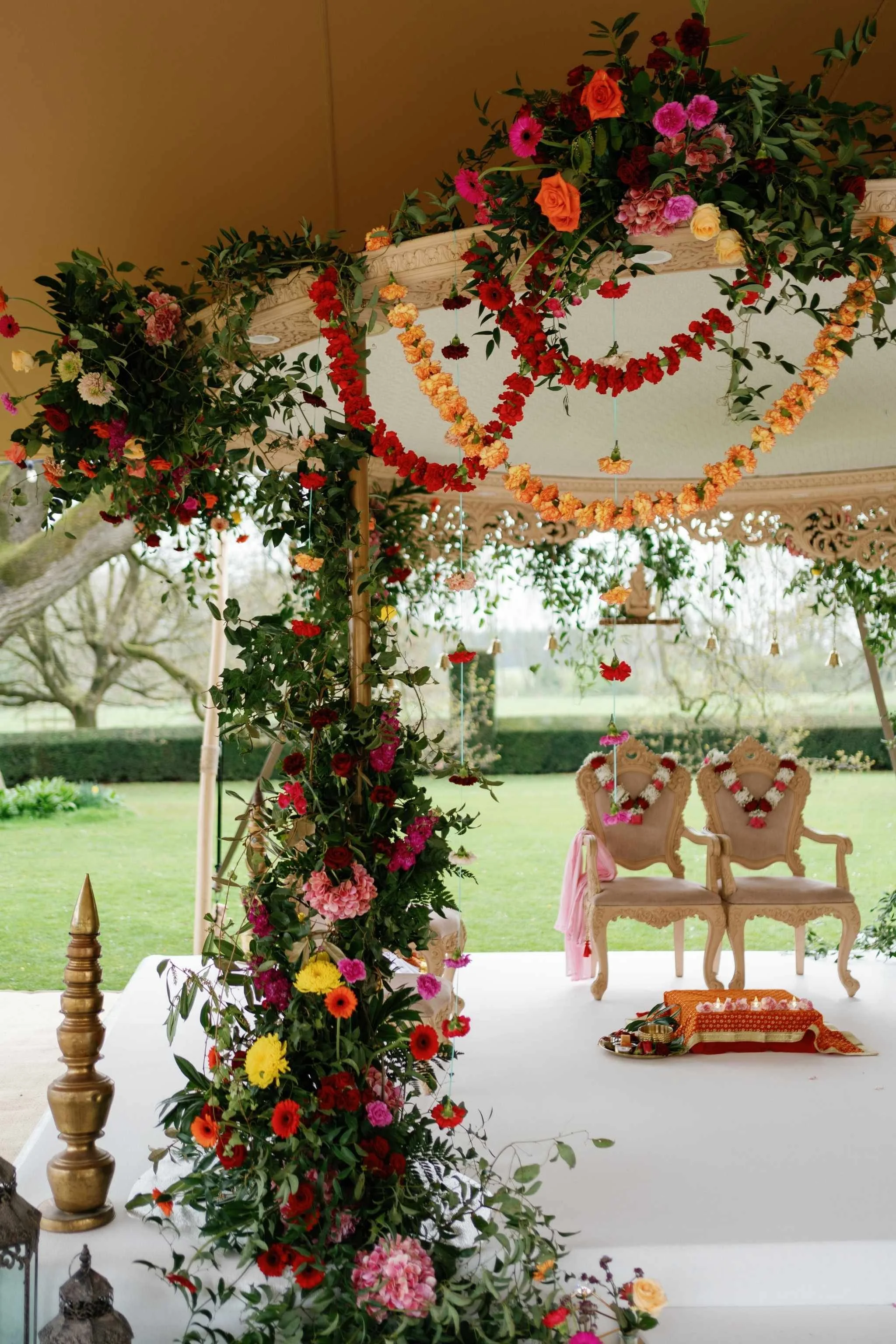 A decorated outdoor venue with floral arrangements, garlands of red, orange, pink, and yellow flowers, and two elegant chairs with floral garlands on the backrests, set on a white platform under a canopy overlooking a green landscape.