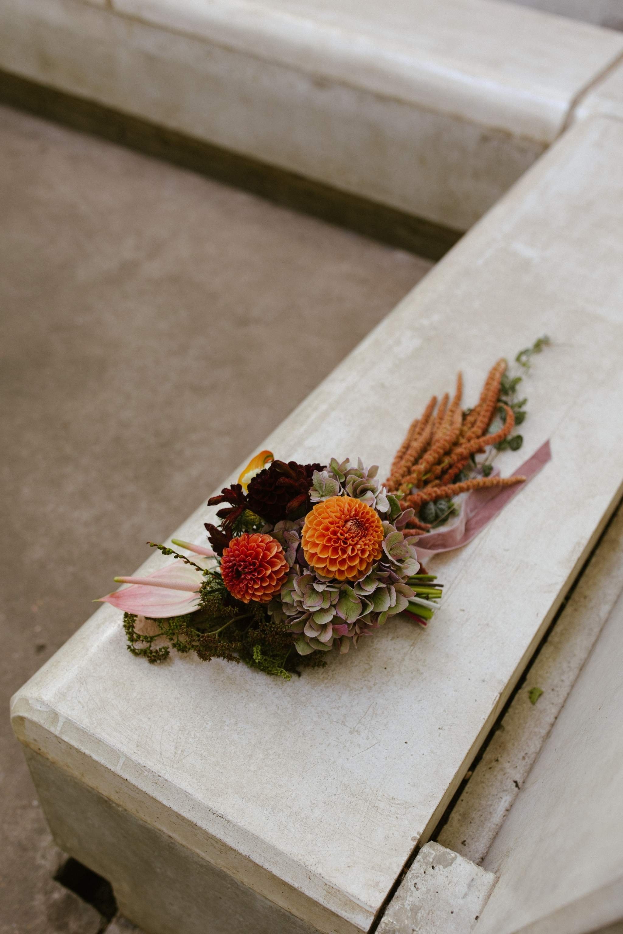 A small bouquet of mixed flowers including orange dahlias, purple and white hydrangeas, and pink anthuriums, wrapped in pink paper, resting on a stone bench.