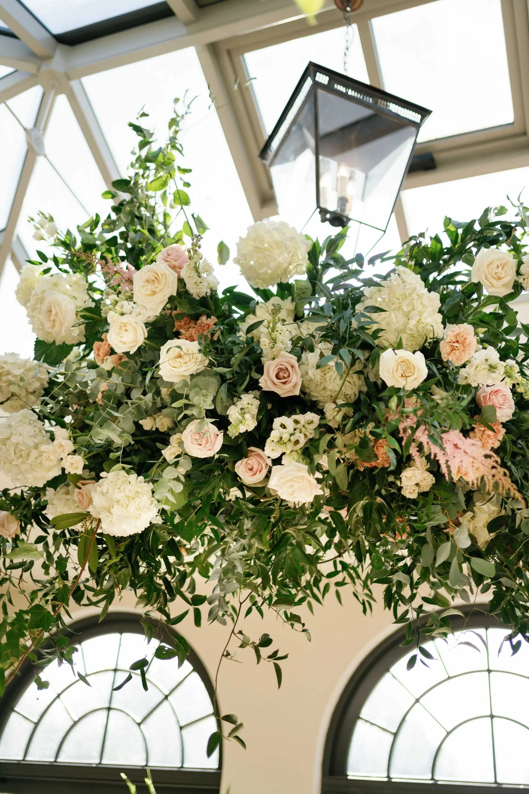 An indoor floral arrangement with white and pale pink roses, white hydrangeas, and green foliage hanging from a ceiling with skylights and arched windows.