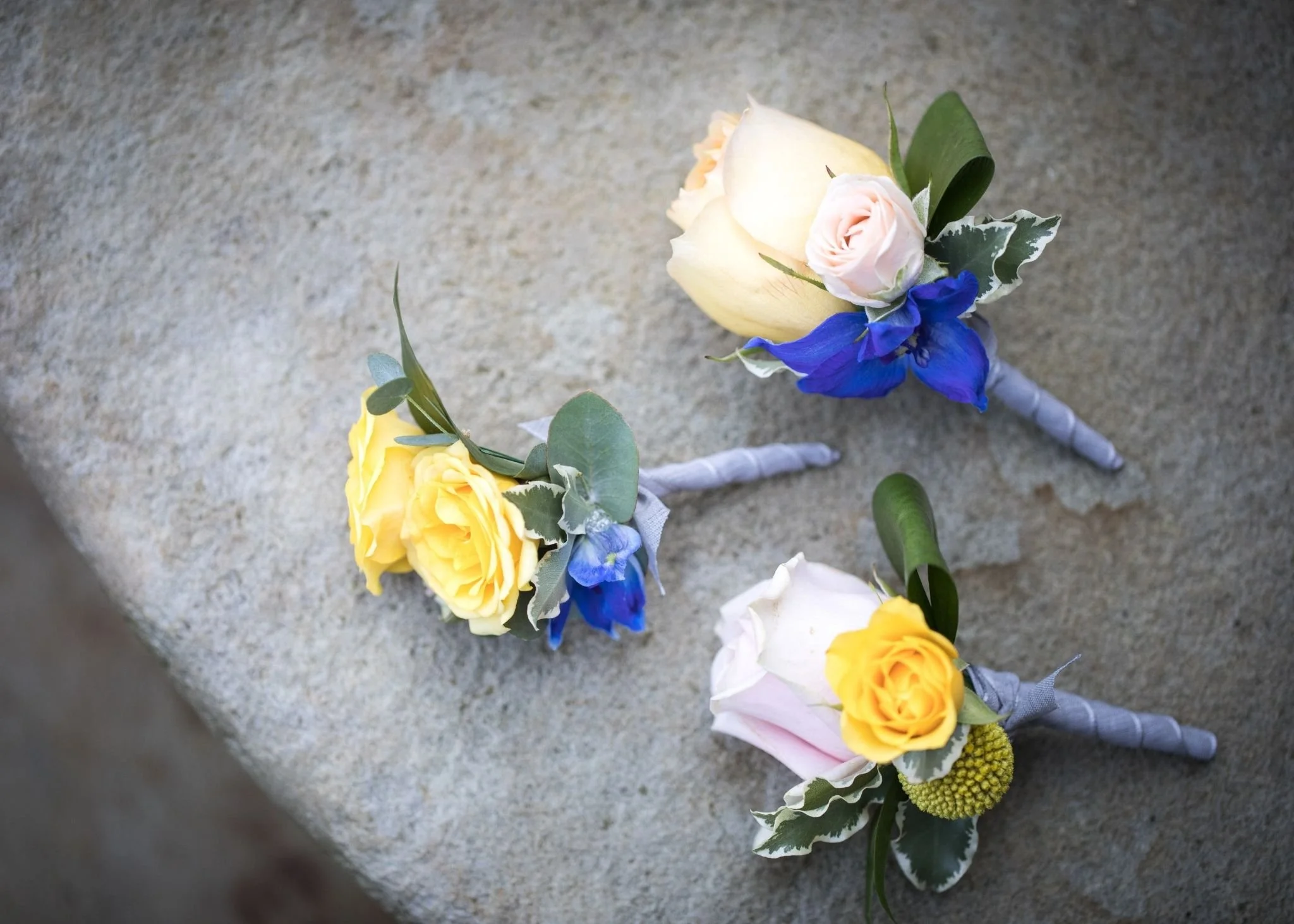 Close-up of a boutonniere with yellow roses, a light pink rose, blue flowers, and greenery, laid on a gray surface.