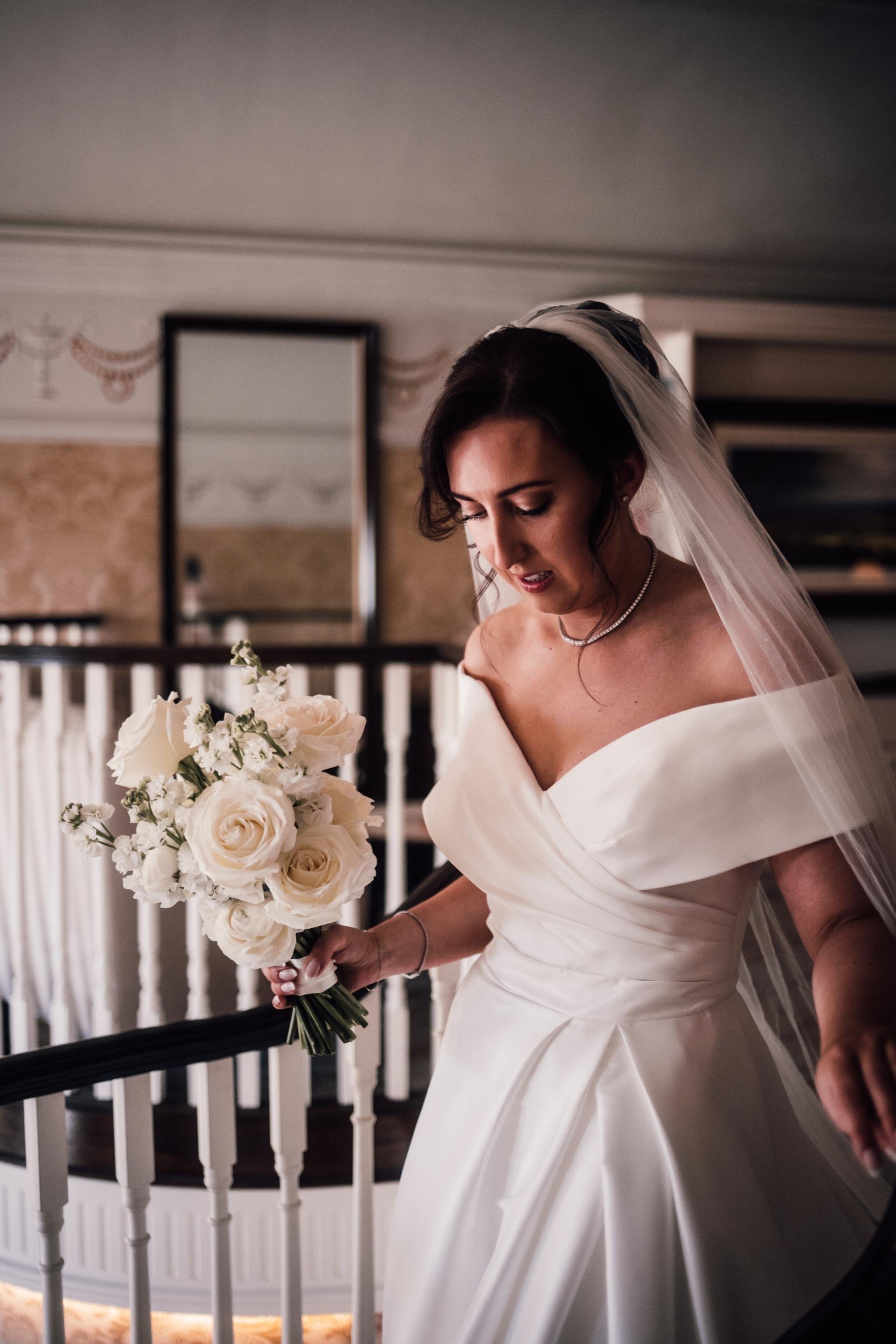 A bride in a white off-the-shoulder wedding gown holding a bouquet of white roses and baby's breath, standing indoors near a staircase with a white railing.