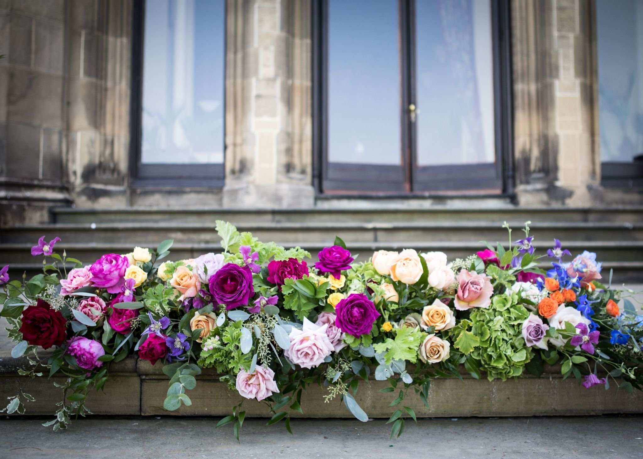Colorful flower arrangement with pink, purple, yellow, and white flowers in front of a stone building with glass door and steps.