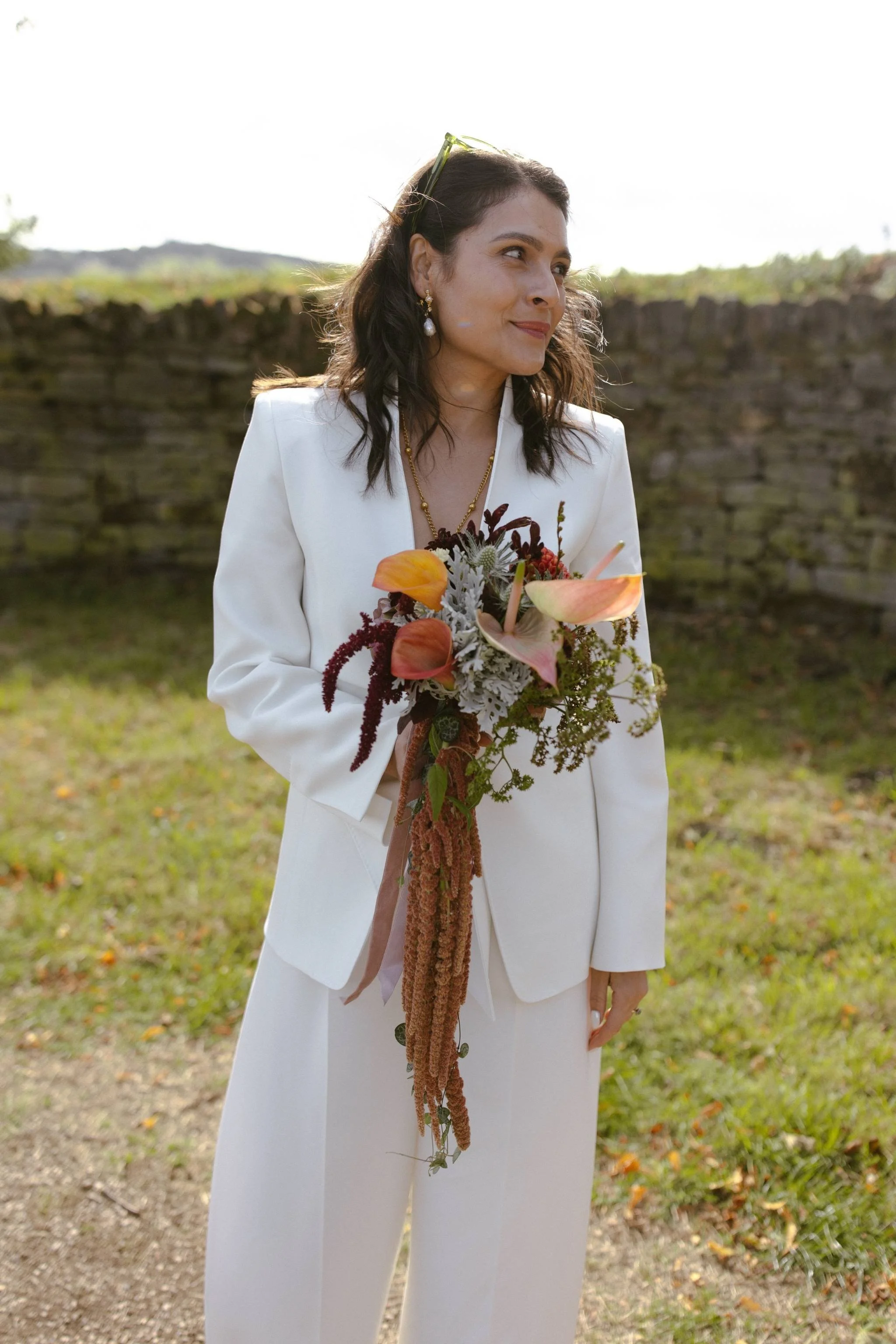 Woman in white suit holding a bouquet of flowers outdoors with a stone wall and green landscape in the background.