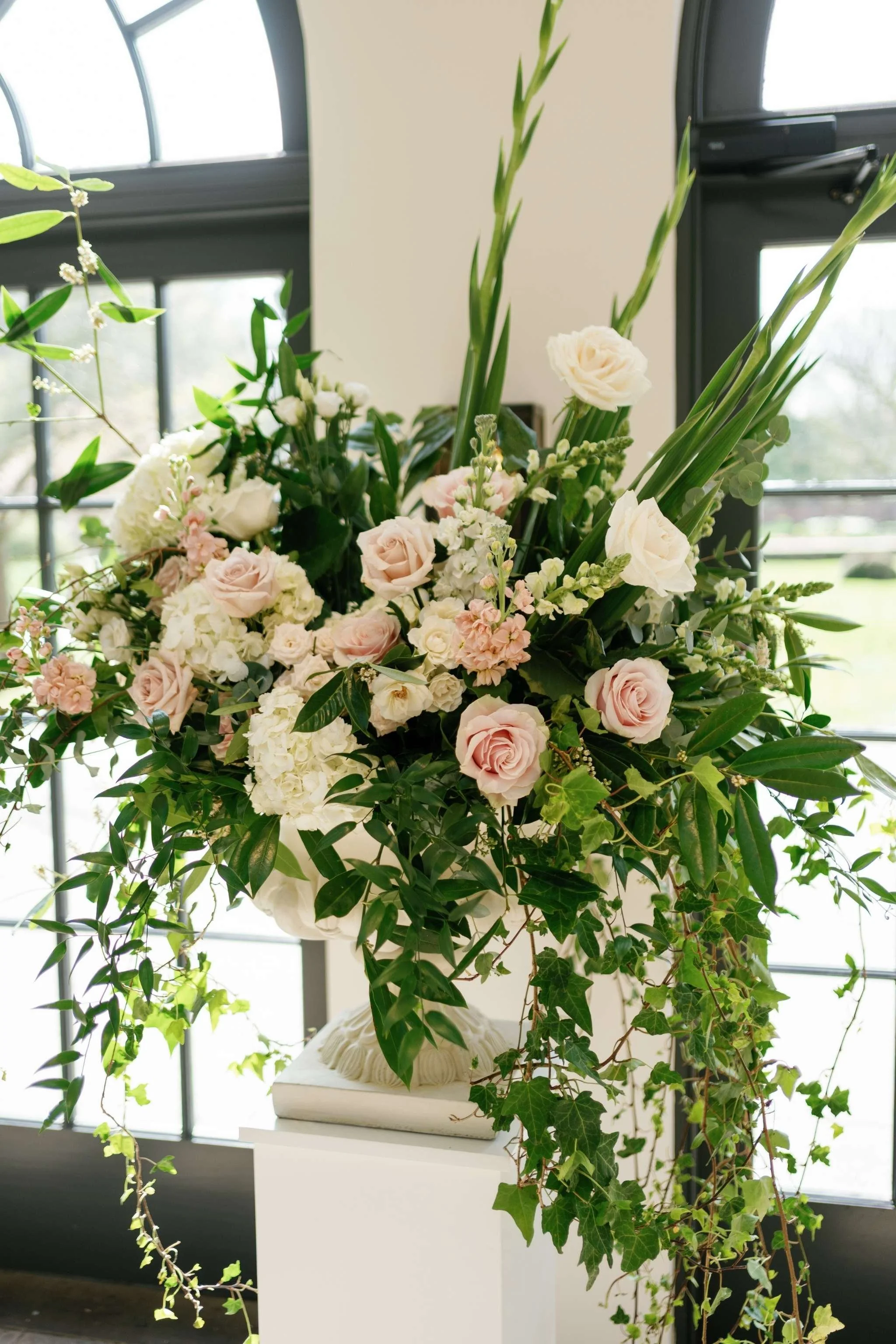 A floral arrangement featuring white roses, hydrangeas, and greenery, placed on a white pedestal with a decorative shell design, in front of large windows letting in natural light.
