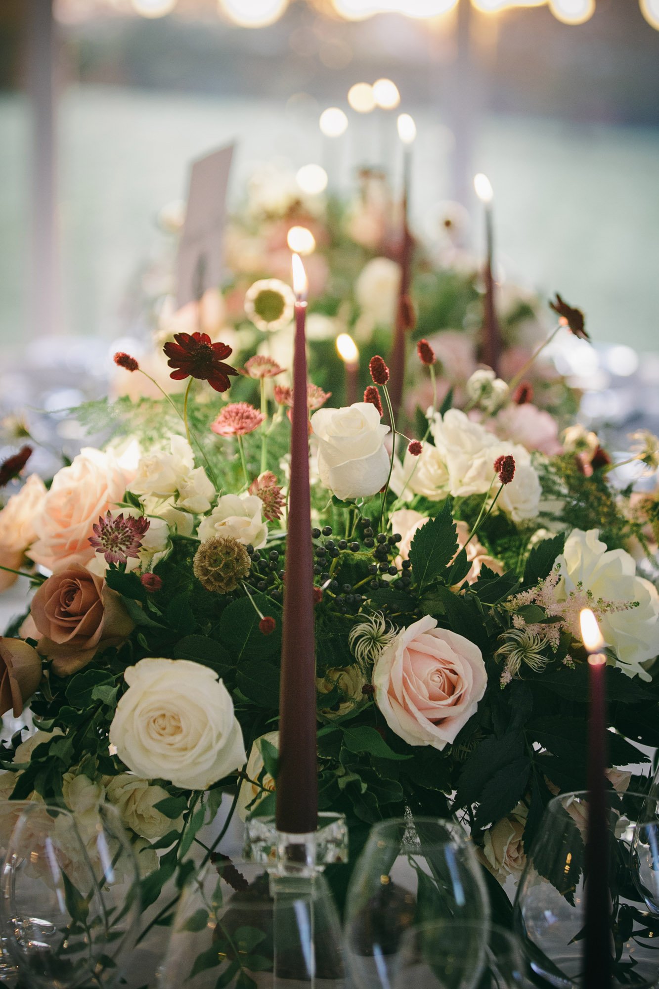 Close-up of a floral centerpiece featuring white and blush roses, greenery, and small flowers with lit purple taper candles, set on a table with wine glasses in a softly lit setting.