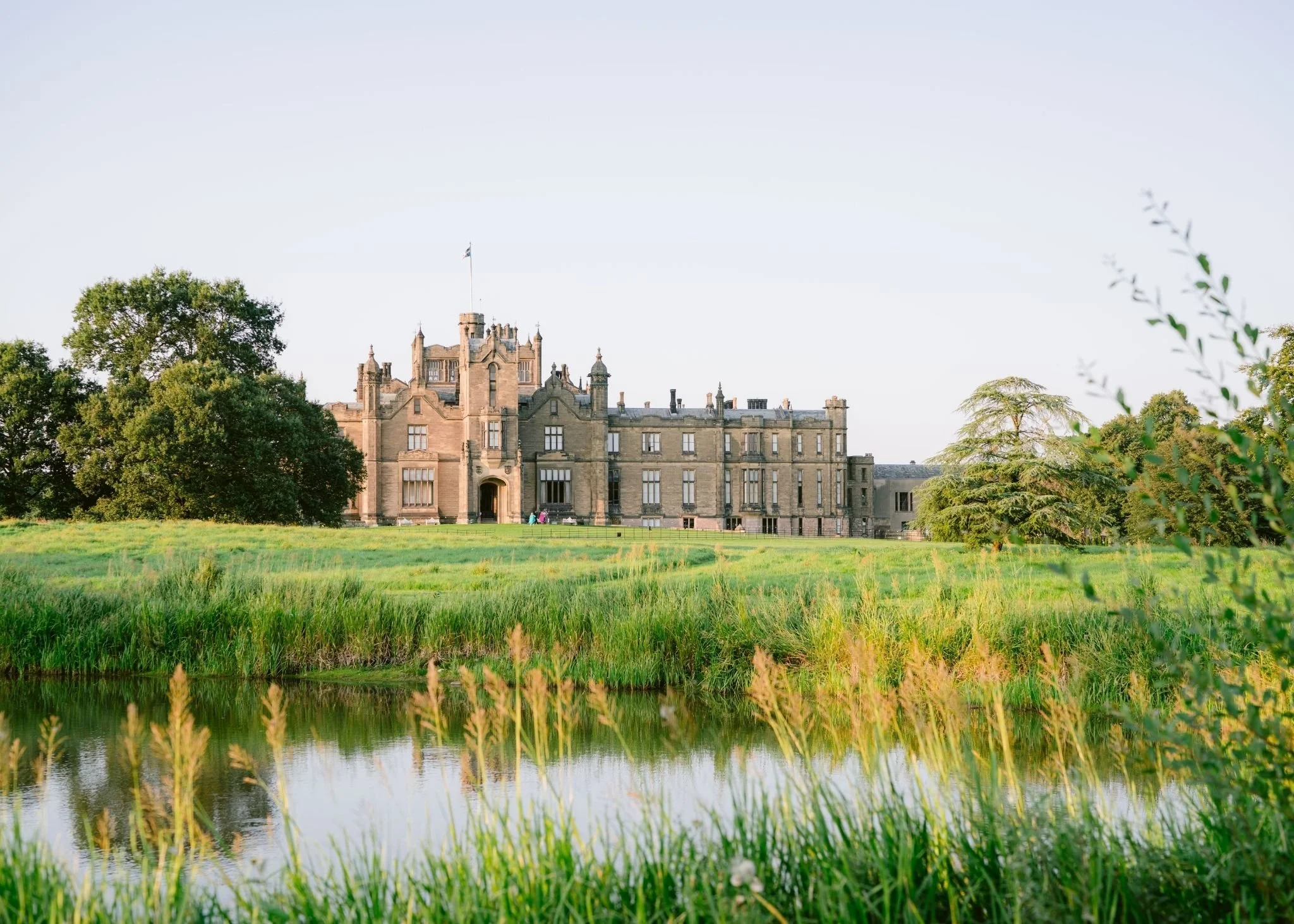 A castle-like building surrounded by greenery and trees, with water in the foreground reflecting the scene on a clear day.
