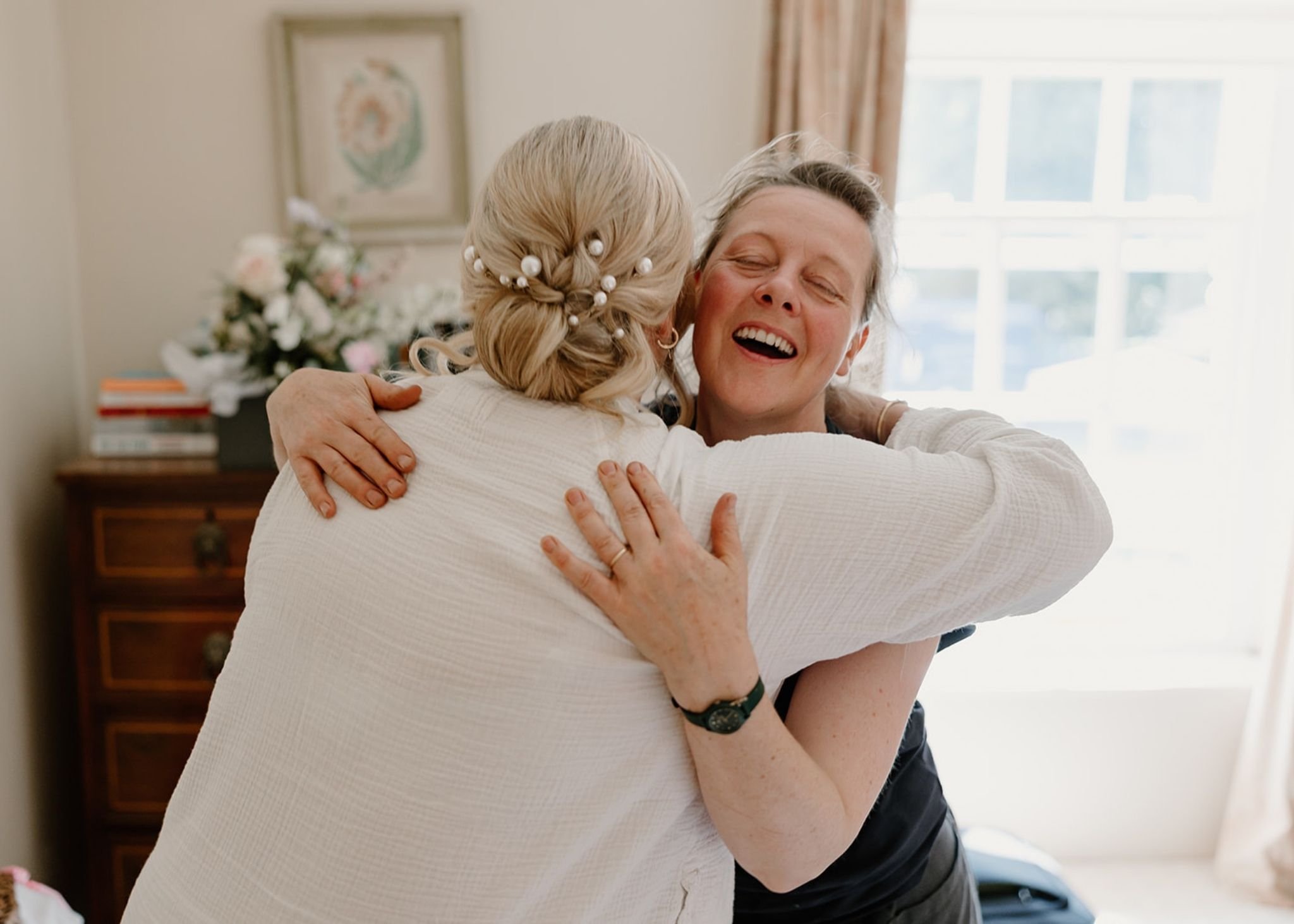 Two women hugging and smiling happily in a room with sunlight coming through the window.