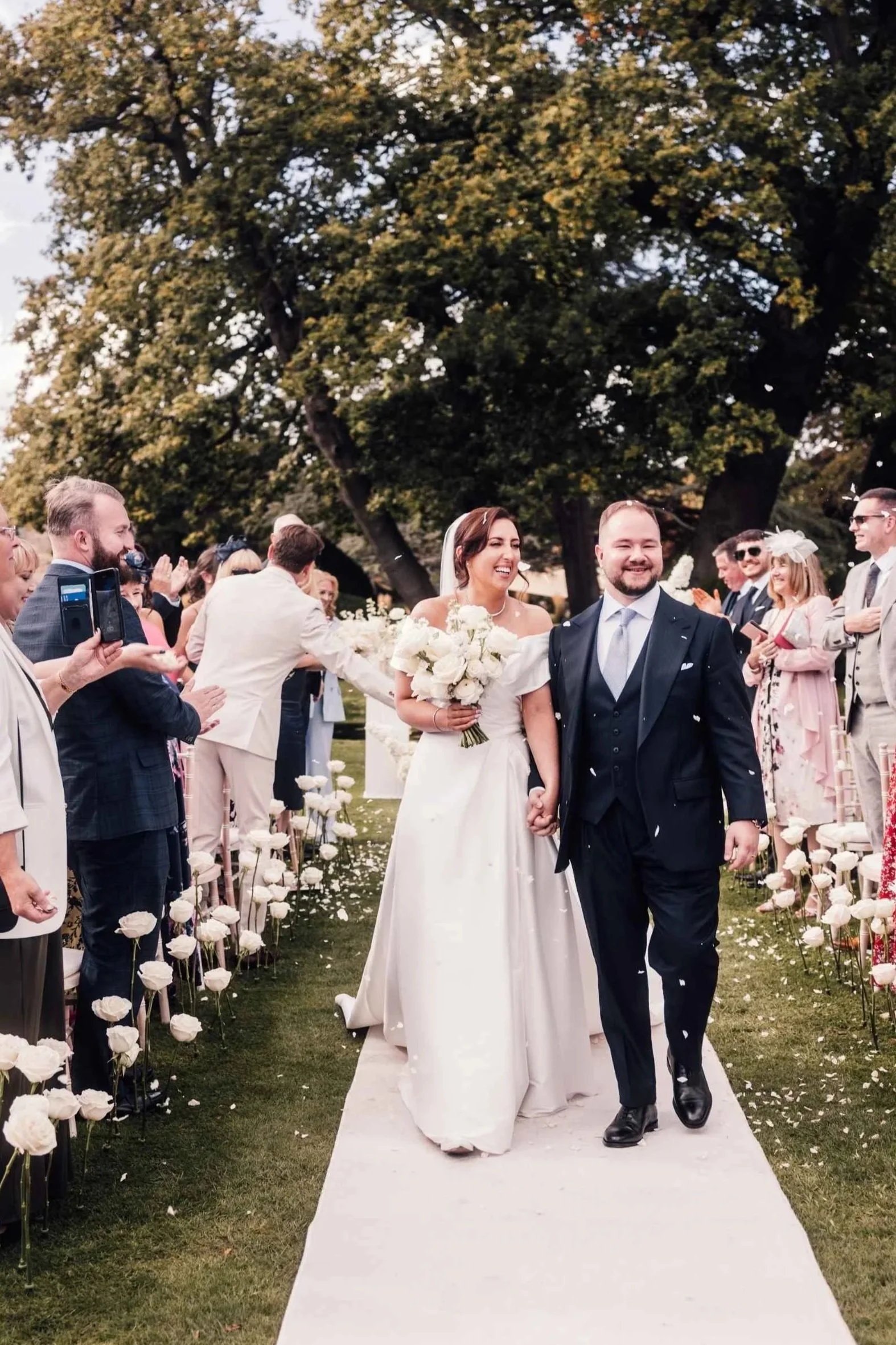 A bride and groom walk hand in hand down an aisle at an outdoor wedding, surrounded by friends and family, with trees in the background and white flower petals on the ground.