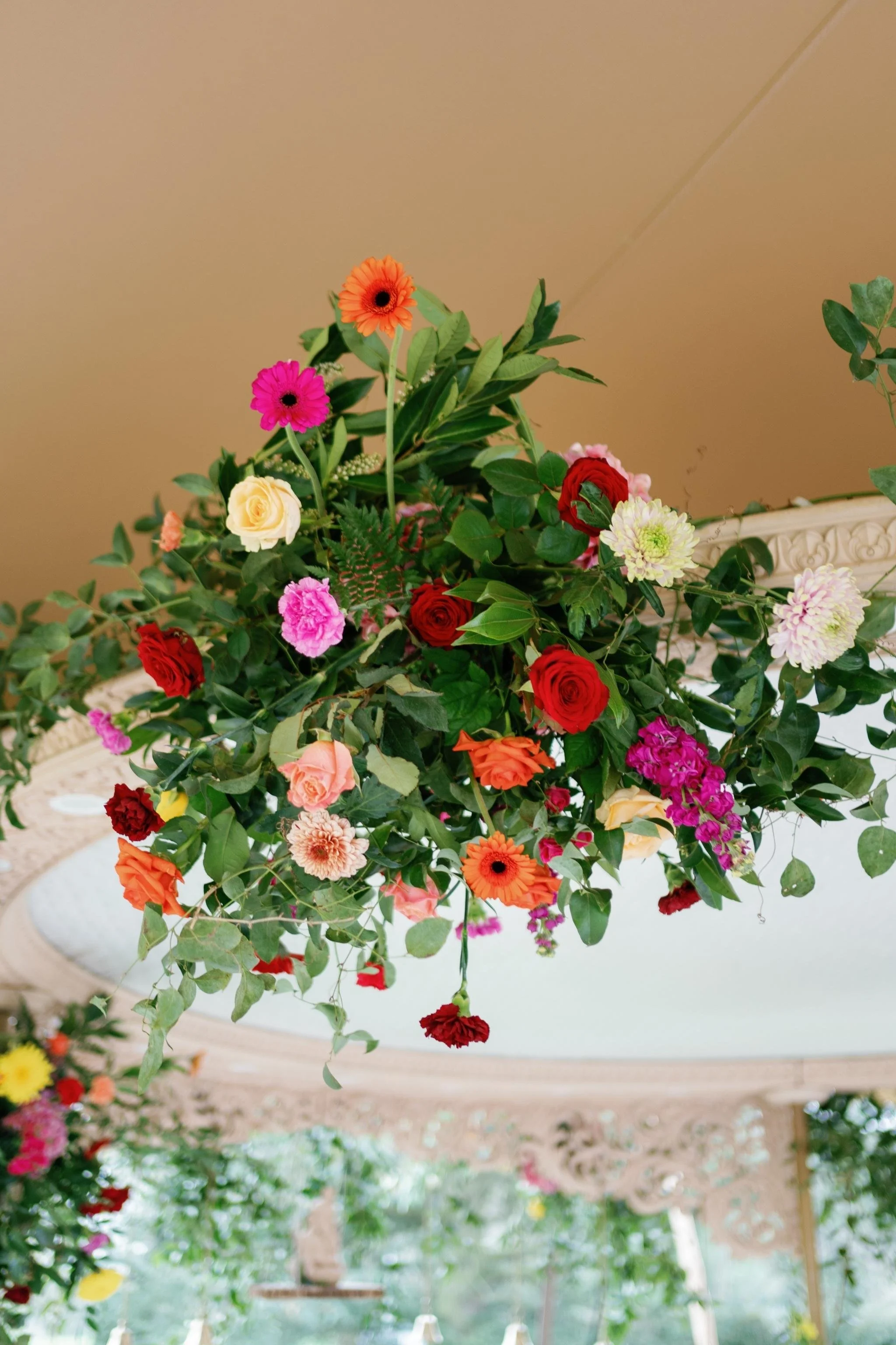 Arrangement of colorful flowers including roses, daisies, carnations, and greenery hanging from a ceiling in a decorated outdoor or garden setting with ornate woodwork.