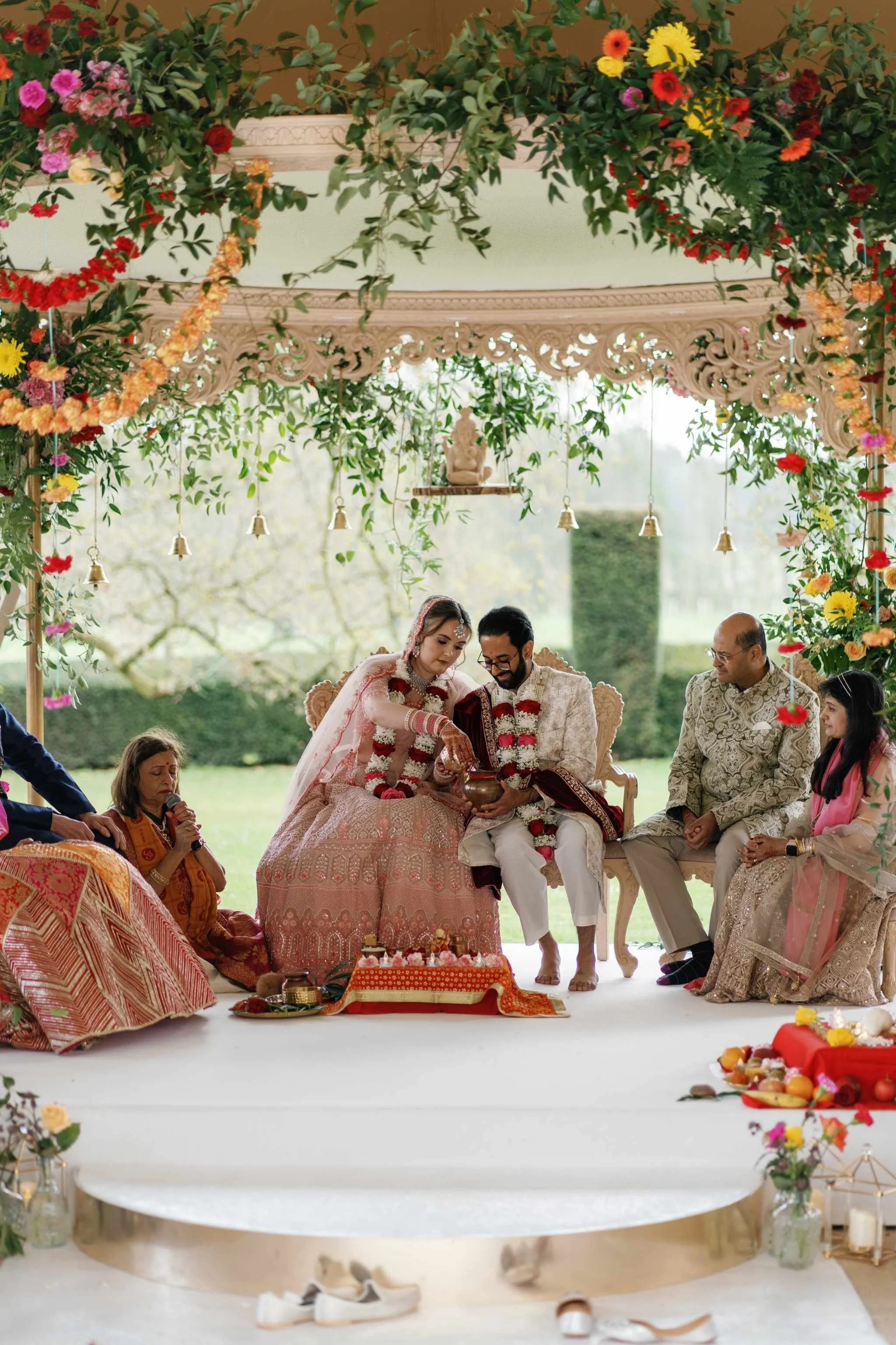 A traditional Indian wedding ceremony with the bride and groom dressed in ornate attire, seated in front of an officiant. The scene is adorned with flowers, greenery, and decorative elements, with family members nearby.