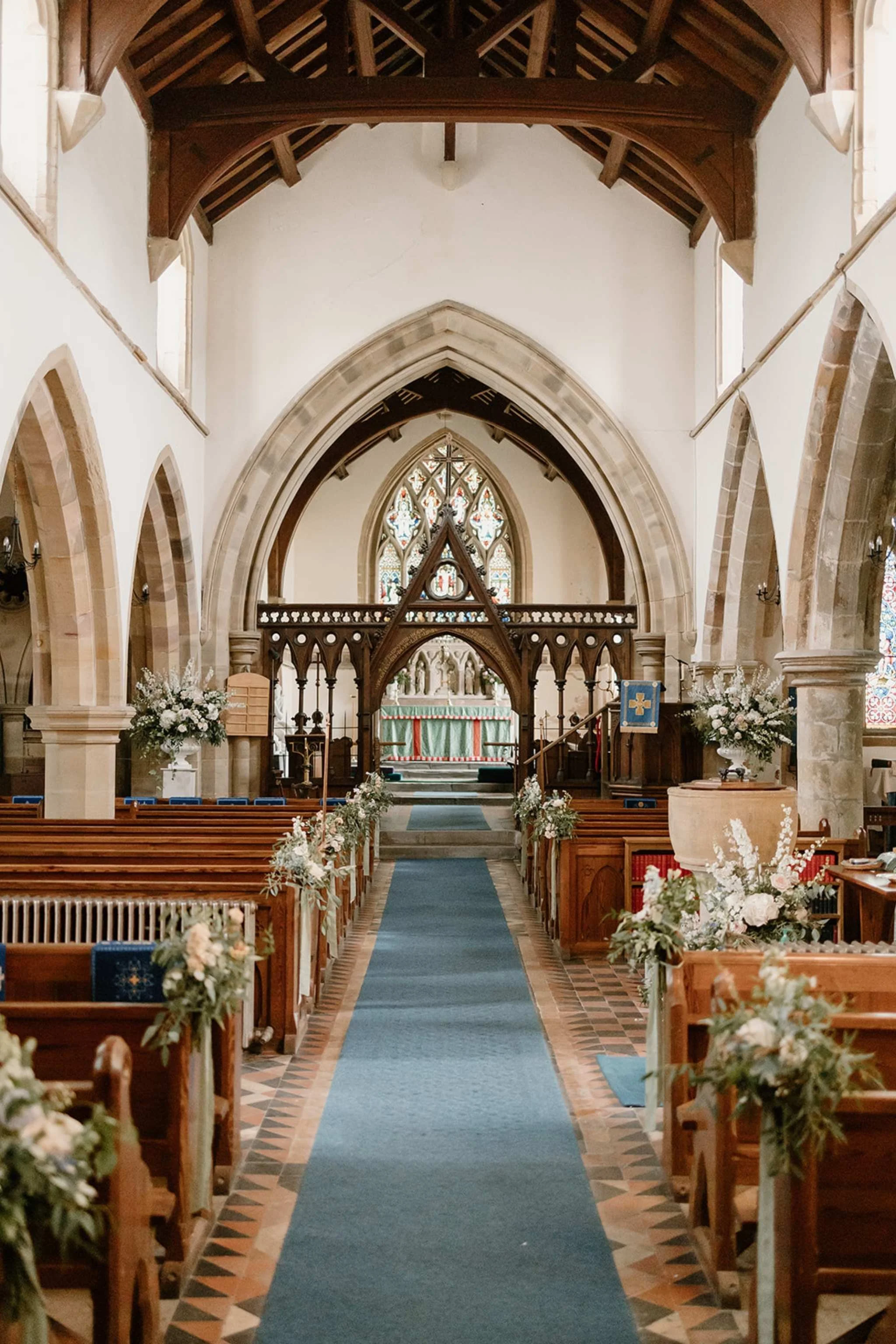 Interior of a church during wedding setup with wooden pews decorated with flowers, a blue aisle runner, stained glass windows, and an altar at the front.