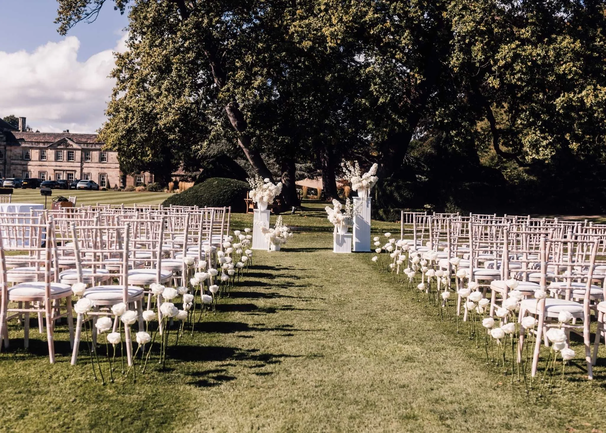 Outdoor wedding setup with white chairs arranged on both sides of an aisle on grass, decorated with white flowers, under large trees with a historic building in the background.