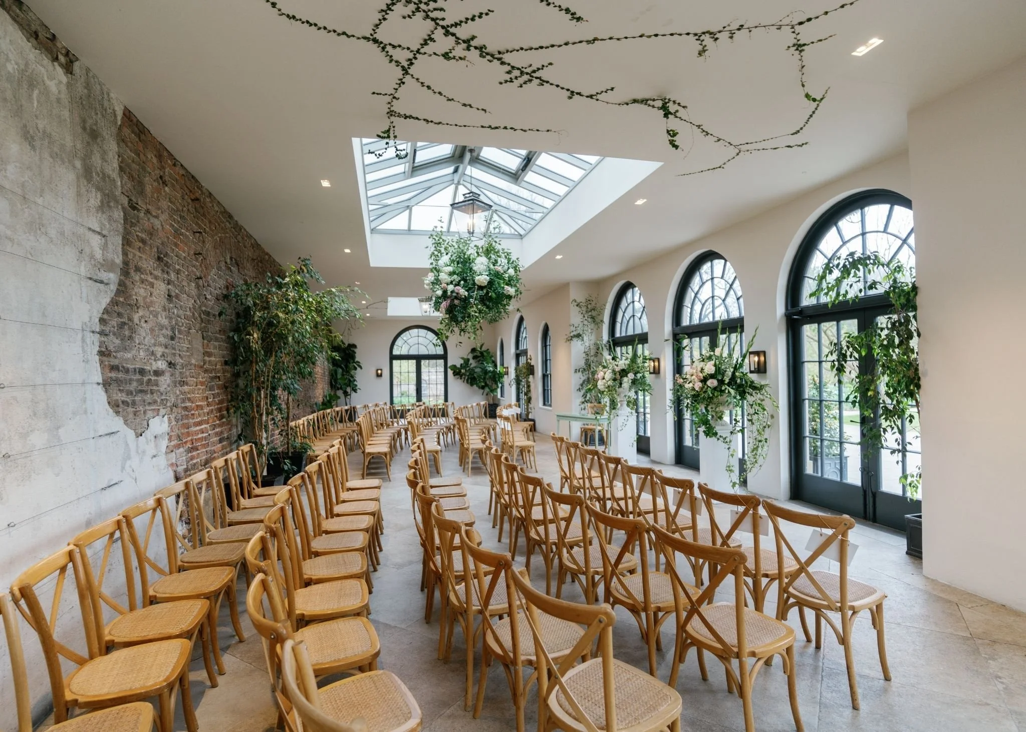 Indoor event space with rows of wooden chairs, large windows, and a glass ceiling letting in natural light. Decor includes hanging flowers and greenery.