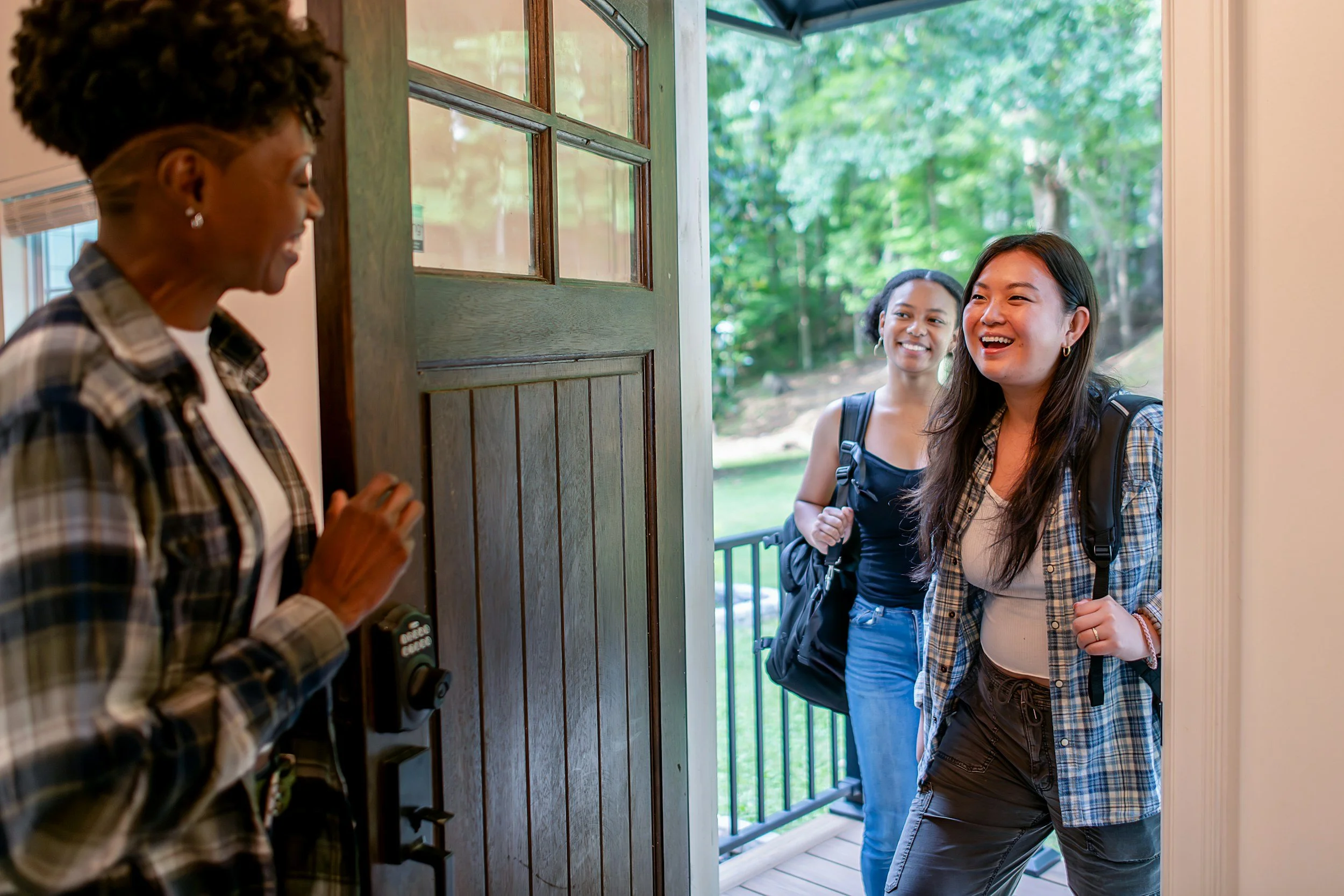 Two women stand at the front door of a house, greeting a visitor inside. The visitor is a woman wearing a plaid shirt, with short curly hair, smiling and holding the door open. The two women at the door are smiling, one with long dark hair, wearing a plaid shirt and backpack, and the other with black hair, wearing a sleeveless top and backpack, standing on a porch with a green, leafy background.