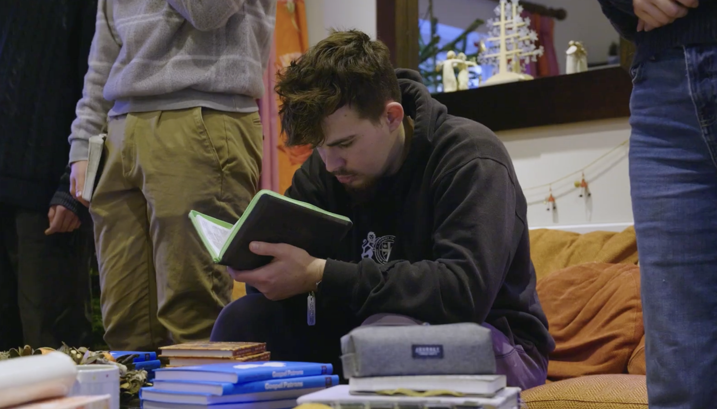 A young man with dark hair and a mustache, sitting on an orange couch, is deeply focused on reading a black book with a green border. Several books and papers are spread out on the table in front of him, surrounded by two standing individuals, one wearing khaki pants and a gray hoodie, and the other in blue jeans.