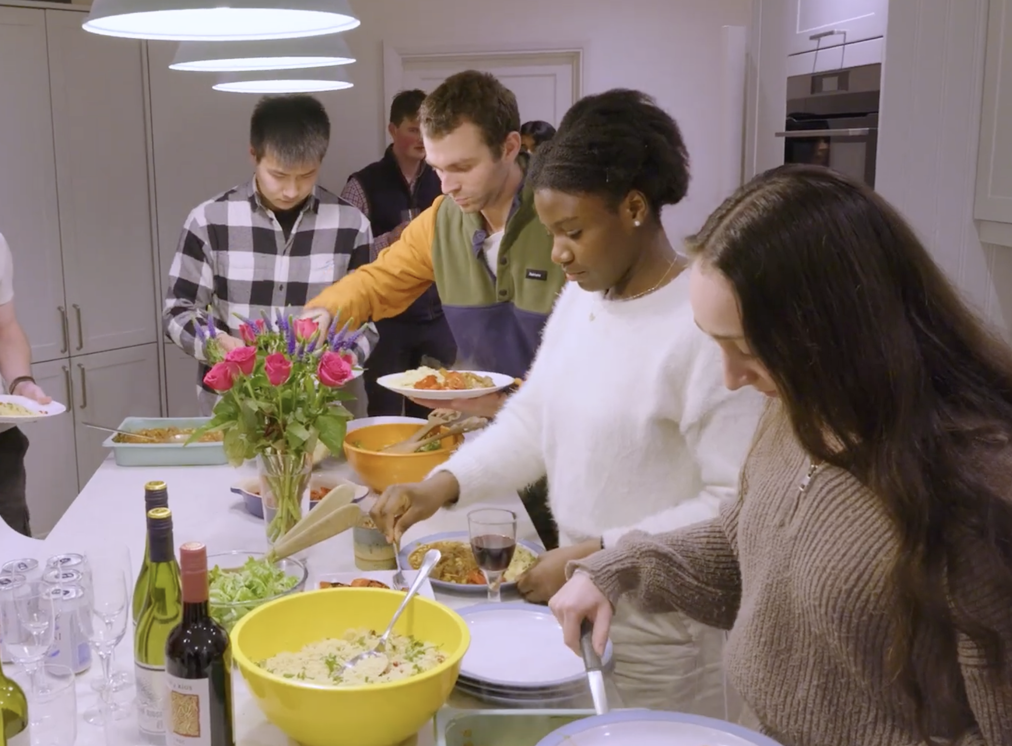 People serving themselves food at a buffet table in a social gathering, with wine bottles and a bouquet of pink flowers on the table.