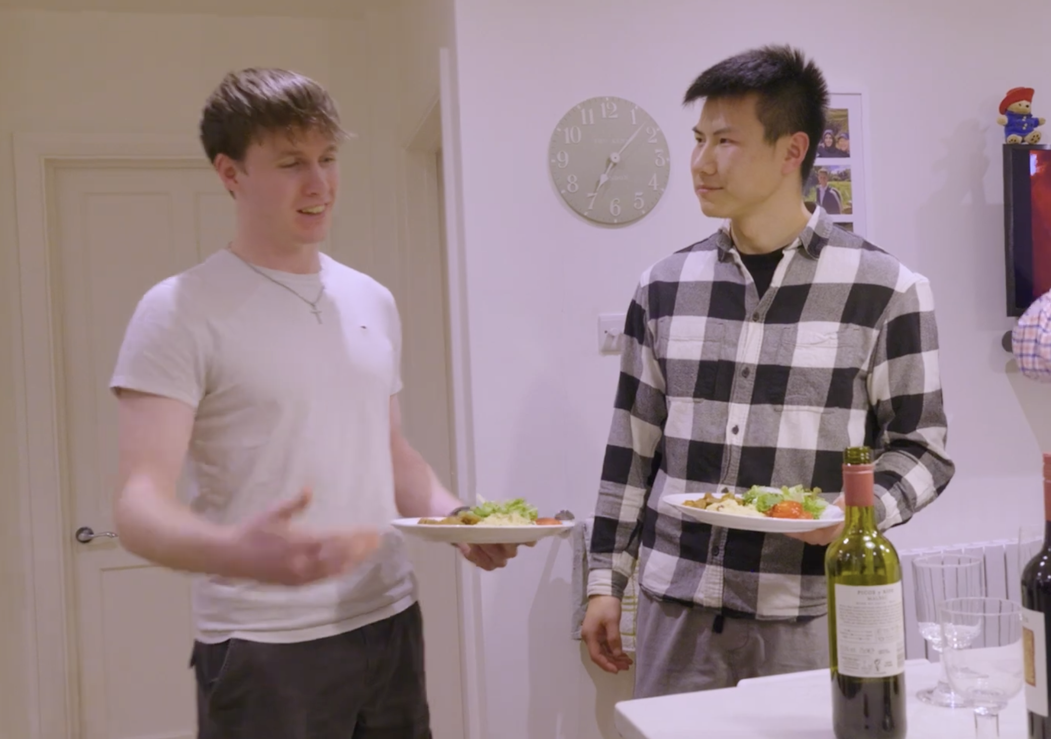 Two young men standing in a kitchen holding plates with salad, engaging in conversation. One is wearing a white t-shirt with a necklace, the other a black-and-white checkered shirt. A bottle of wine and glasses are on the table.