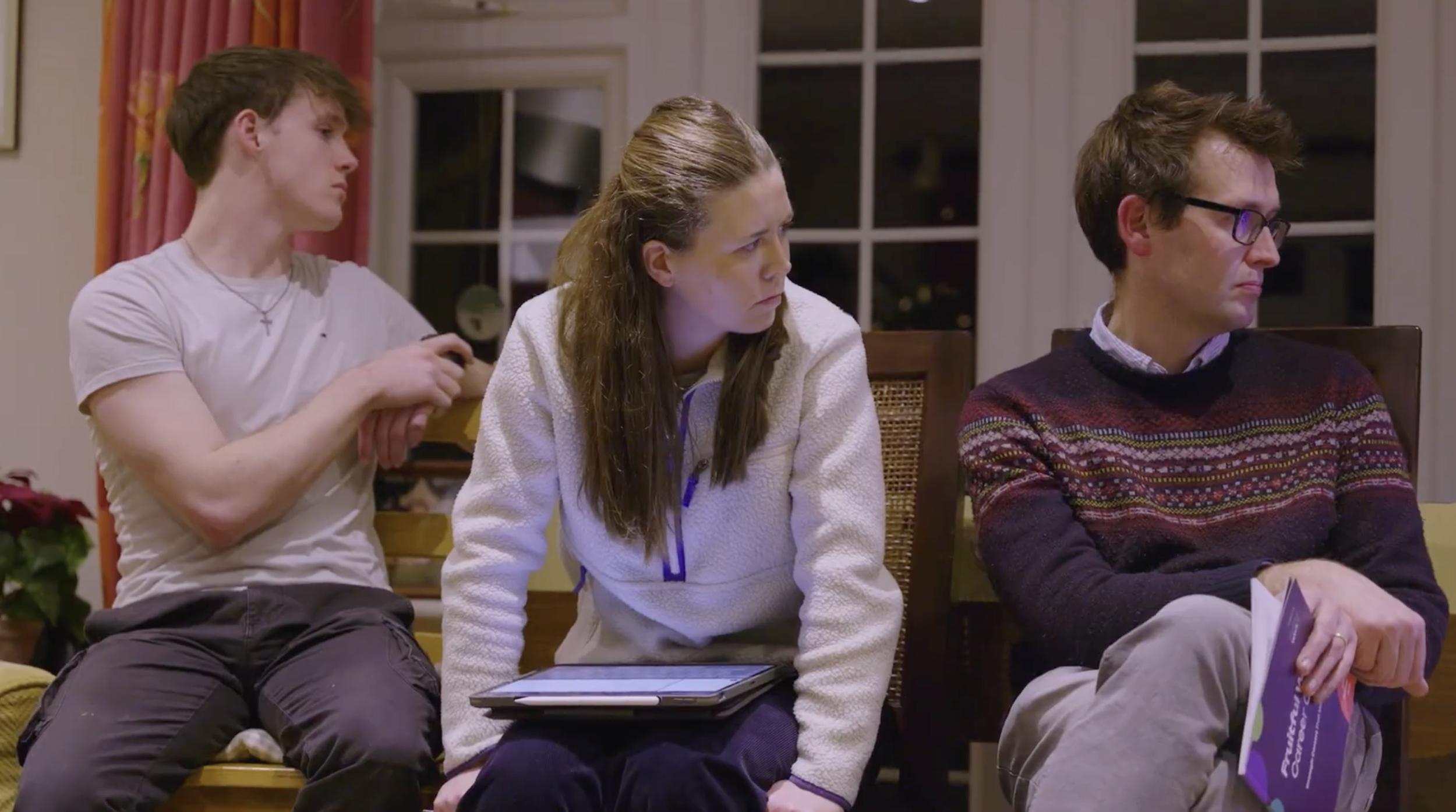 Three people sitting on chairs indoors, engaging in a meeting or event, with a woman in the center looking to her left, a man on the left holding a phone, and an older man on the right holding a pamphlet.