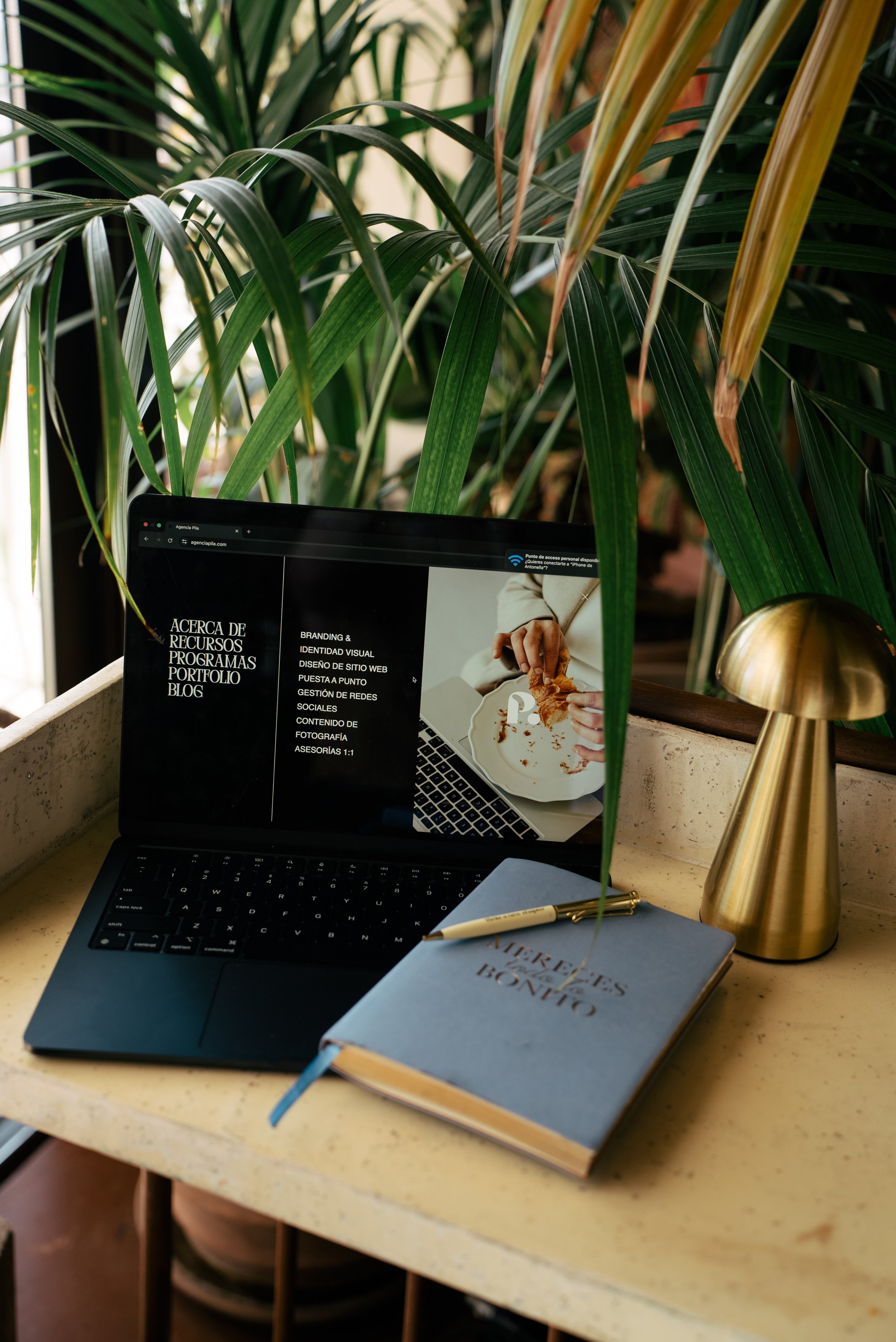 Estación de trabajo en un escritorio con una laptop, libreta azul con pluma dorada, una lámpara dorada, y plantas verdes de fondo.