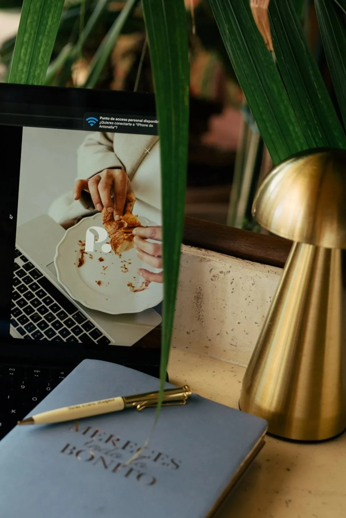 Mostrando una laptop con una foto de una mano comiendo un pastel. En primer plano, hay una libreta azul con un bolígrafo dorado, y una lámpara de superficie dorada. La escena está decorada con plantas verdes.
