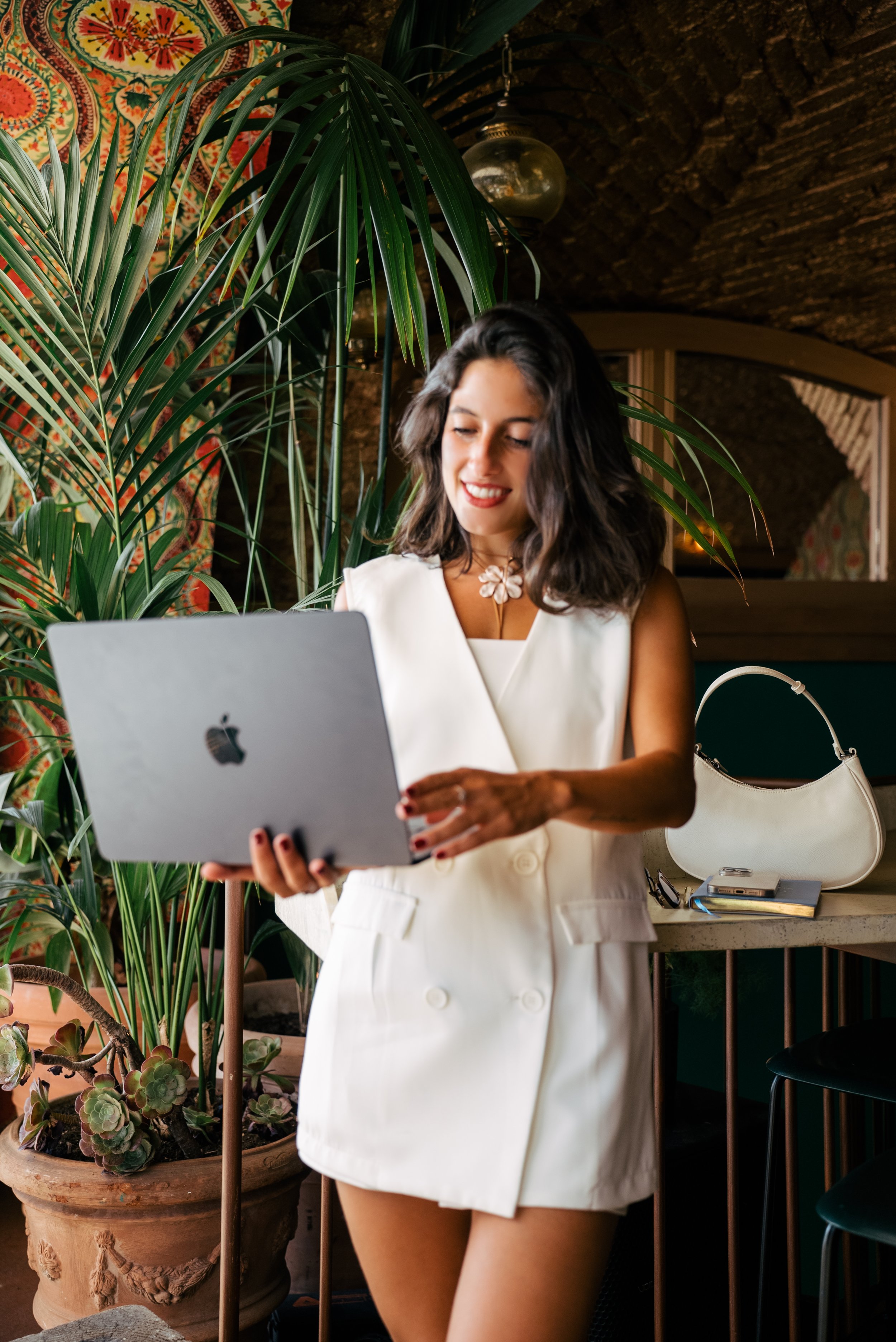Mujer sonriente con vestido blanco sosteniendo una computadora portátil en un espacio con plantas verdes y decoración cálida.