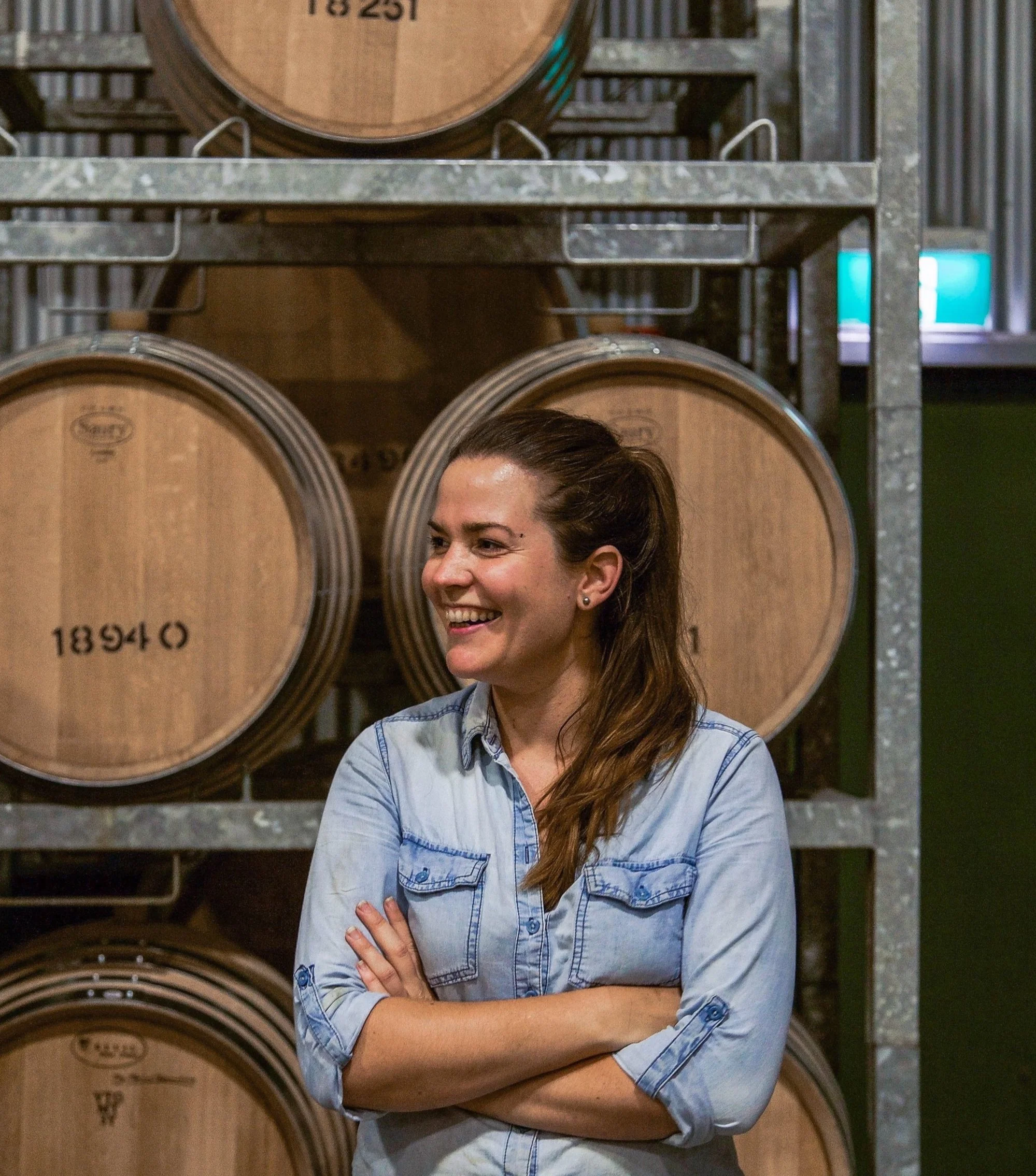 Kate Sturgess - A winemaker with brown hair in a ponytail, wearing a light denim shirt, smiling, standing with arms crossed in front of wine barrels in a cellar.