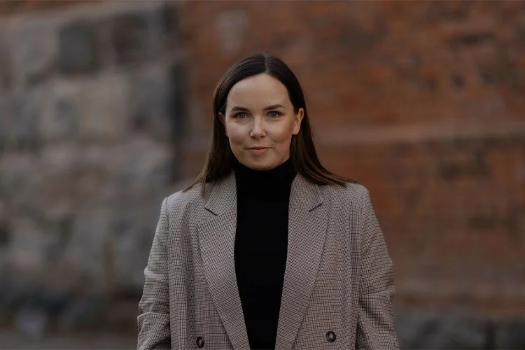 Nainen seisoo ulkona tiiliseinän edessä, pukeutuneena beigeen takkiin ja mustaan puseroon. Henkilö on kiinteistönvälityksen LKV-pätevyyden omaava assistentti Kuopiosta.
